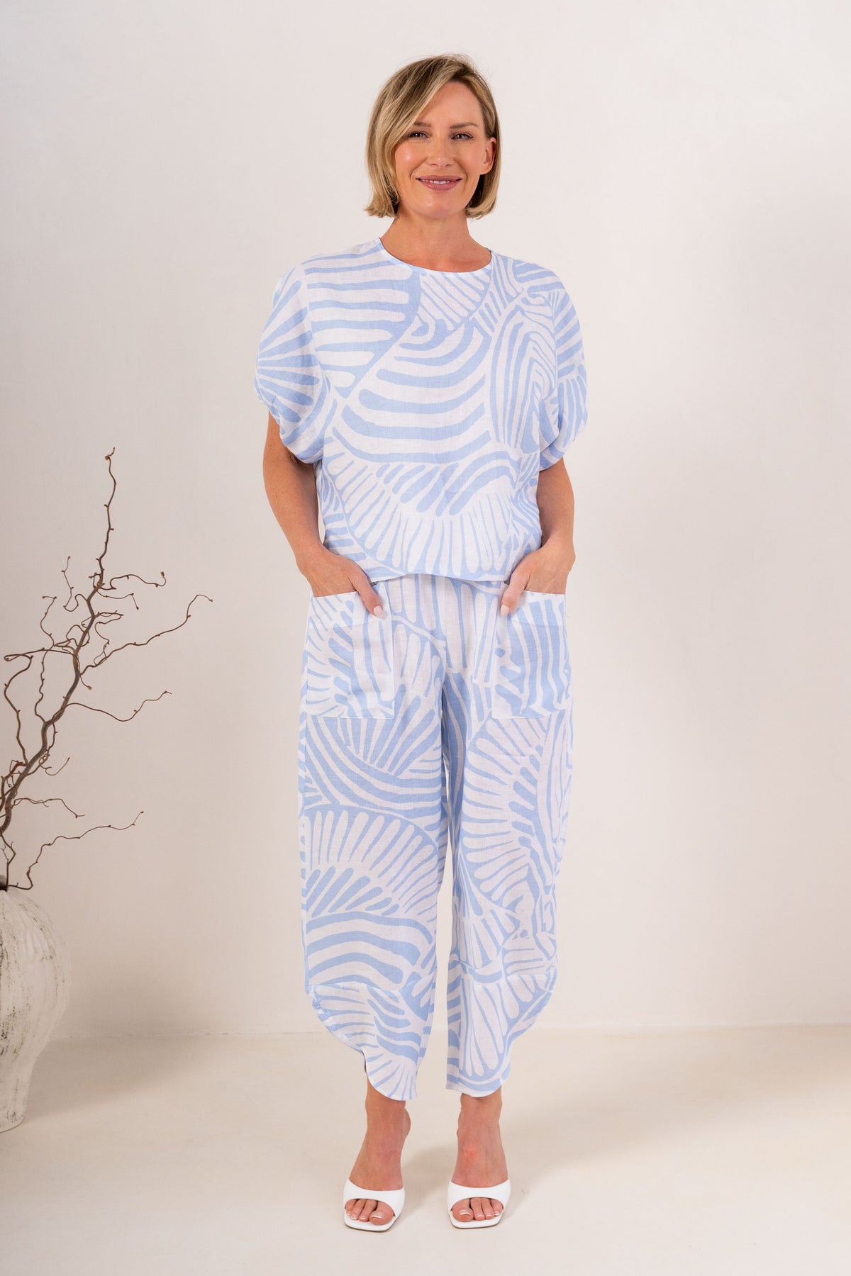 A woman modeling a light blue and white Palm Breze See Saw Linen two-piece set consisting of a short-sleeve top and matching pants, standing against a plain off-white studio background.