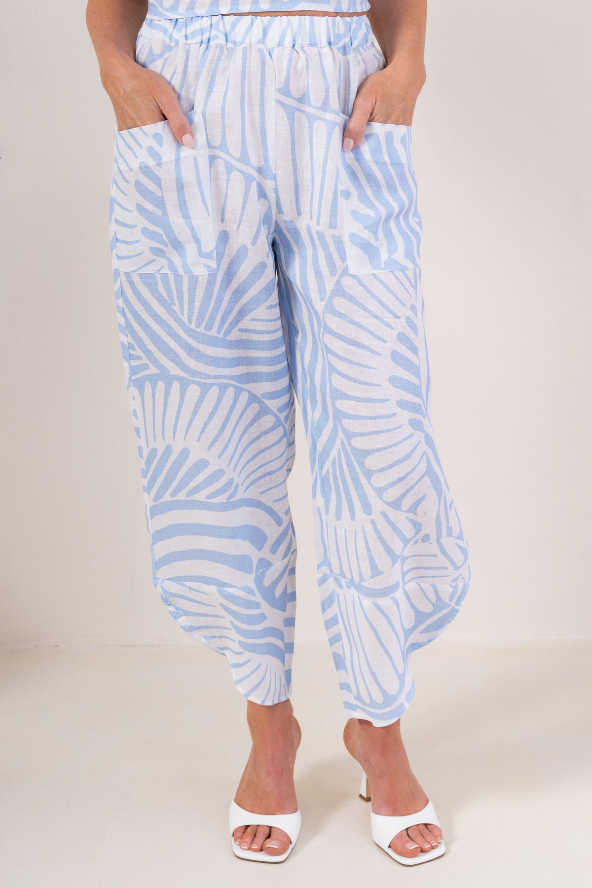 See Saw Palm Breeze print linen See Saw pants with side pockets, shown on a model wearing white heeled sandals against a plain beige background.