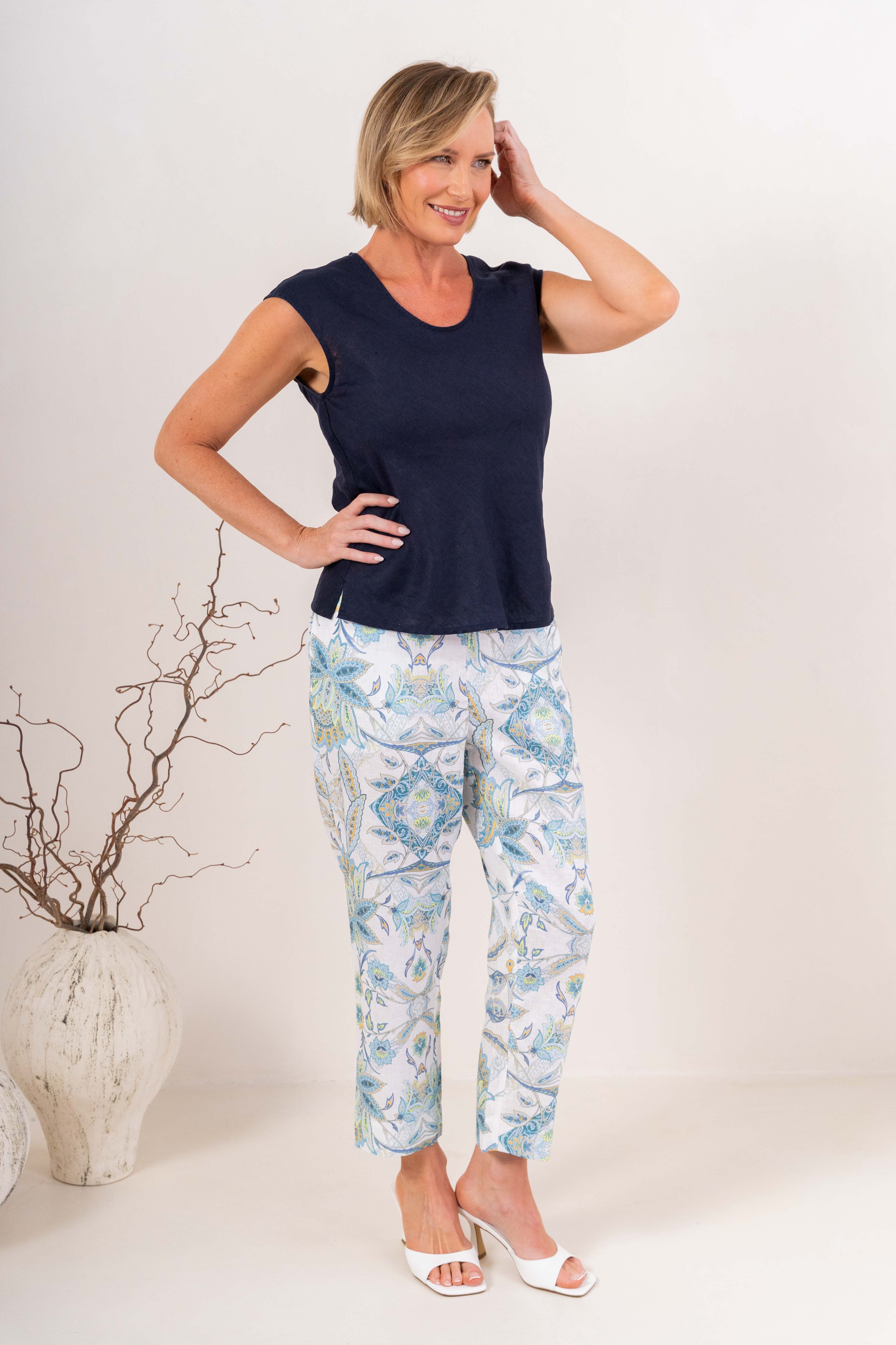 A smiling blonde woman wearing a navy sleeveless V-neck top in linen by See Saw and matching white pants, standing against a plain light background.