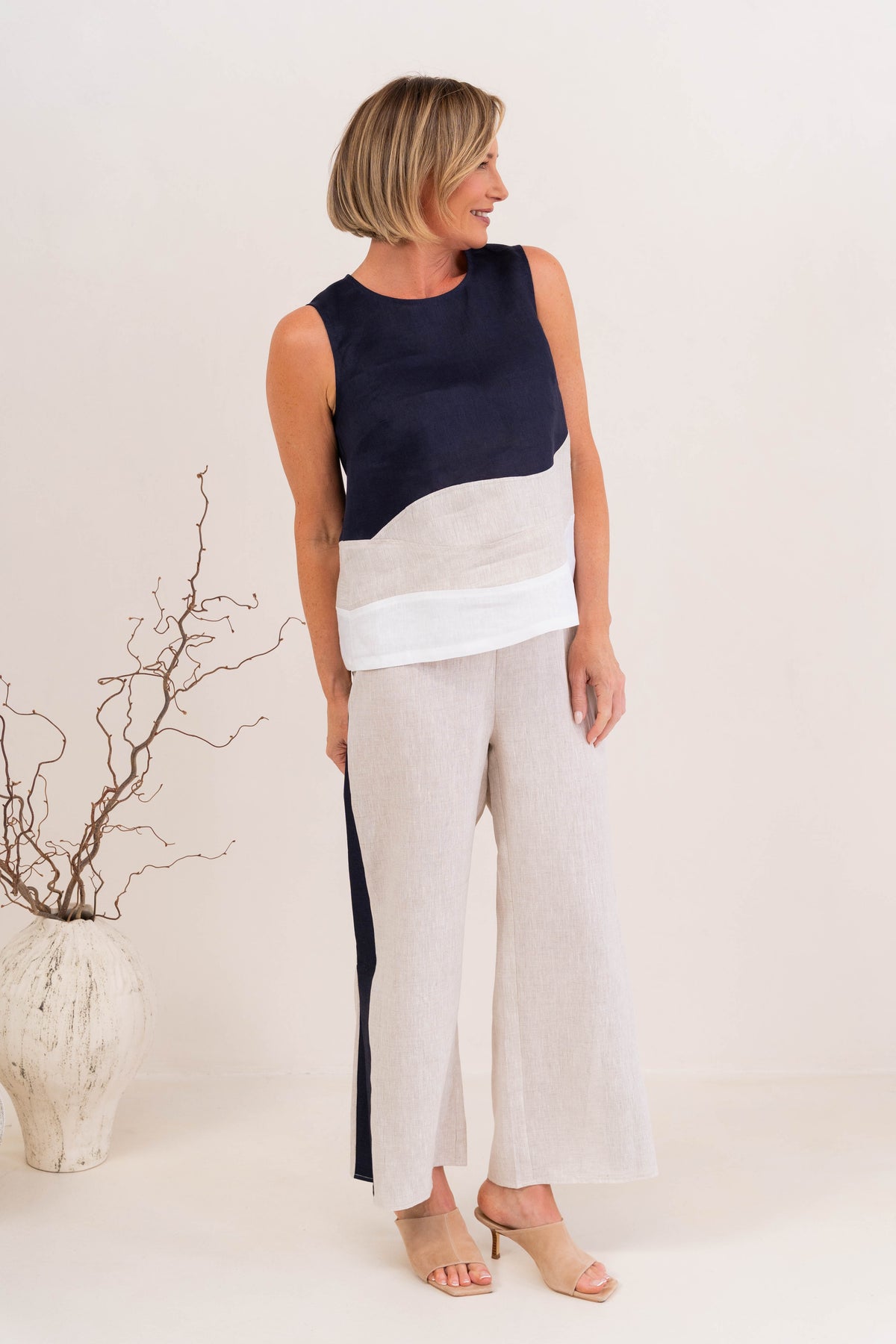 A woman modeling a sleeveless See Saw Linen color-block top and matching wide-leg trousers in navy, off-white, and light beige, standing against a plain light background.