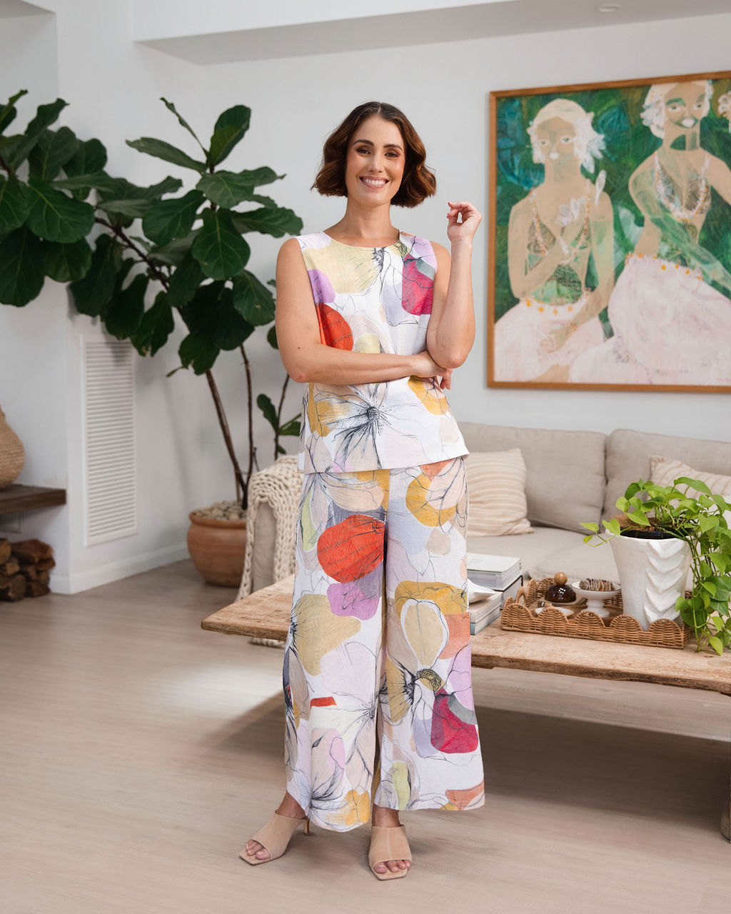A woman modeling a sleeveless Petal Drift linen See Saw Top and pant set with a colorful abstract floral print, standing in a bright, modern living room.