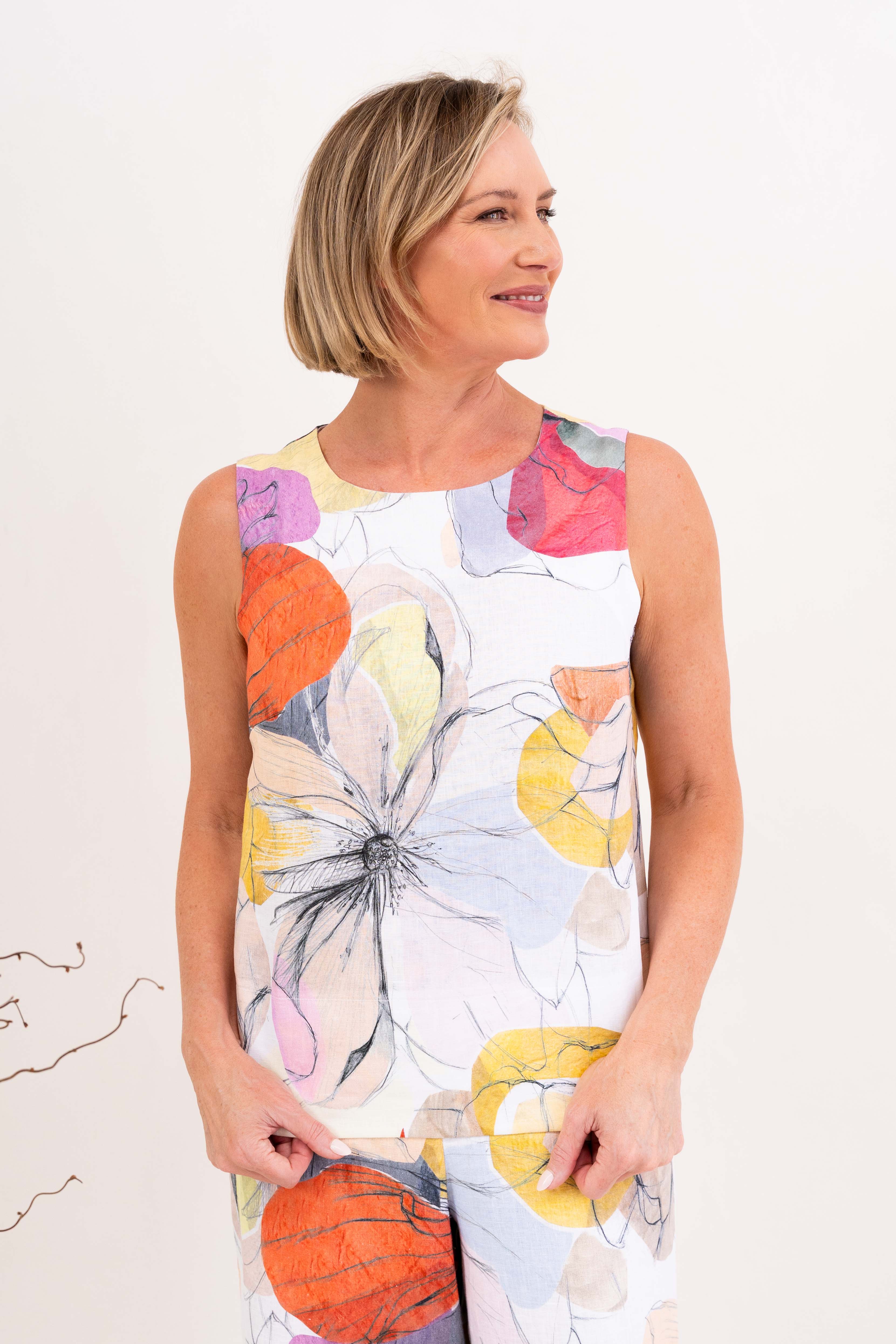 A woman modeling a sleeveless Petal Drift linen See Saw Top and pant set with a colorful abstract floral print, standing in a bright, modern living room.