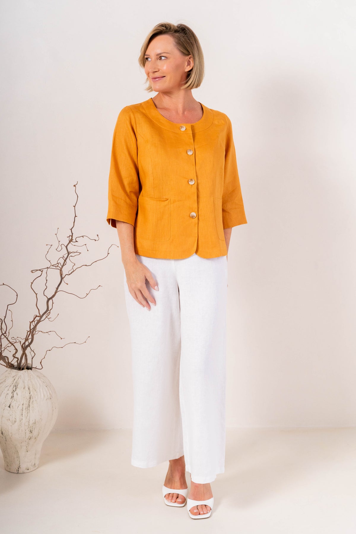 Woman modelling marigold linen see saw utility jacket top and white pants and against a plain background.