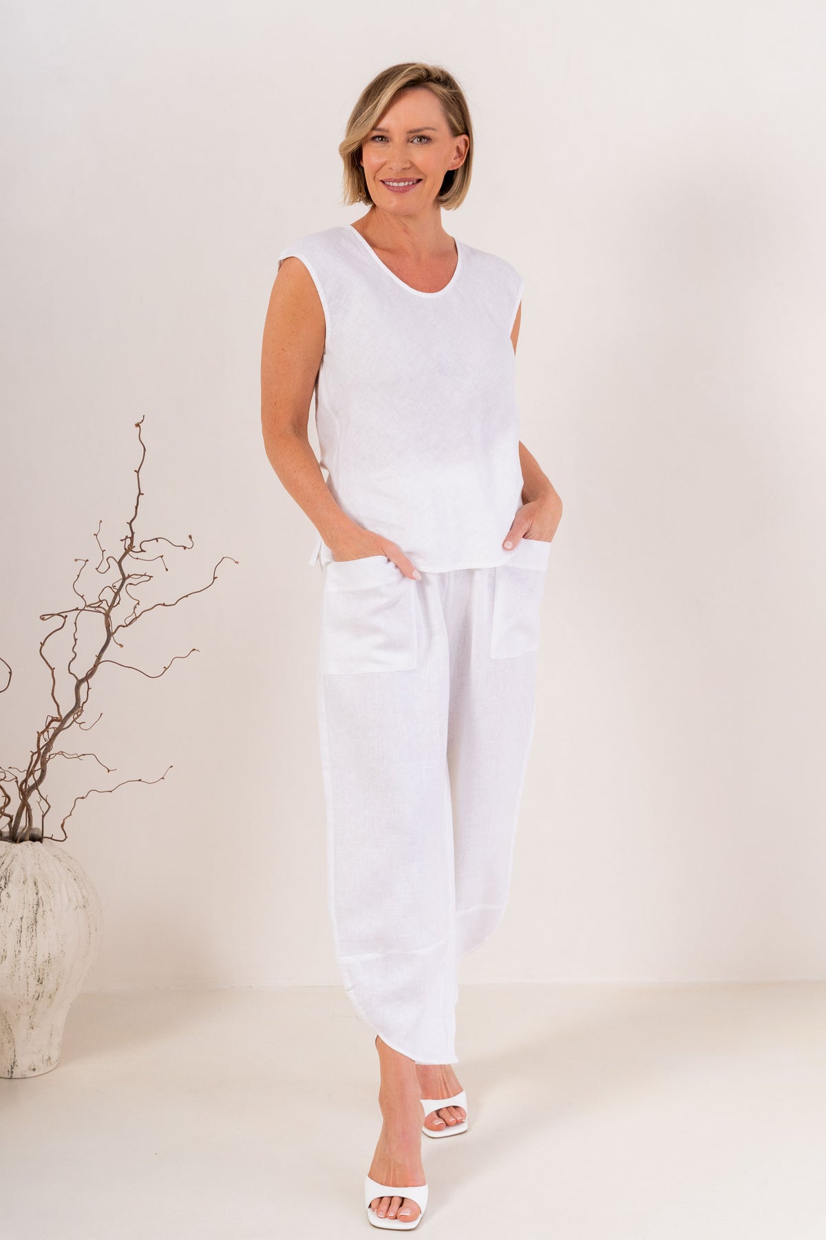 A smiling blonde woman wearing a white sleeveless V-neck top in linen by See Saw and matching white pants, standing against a plain light background.