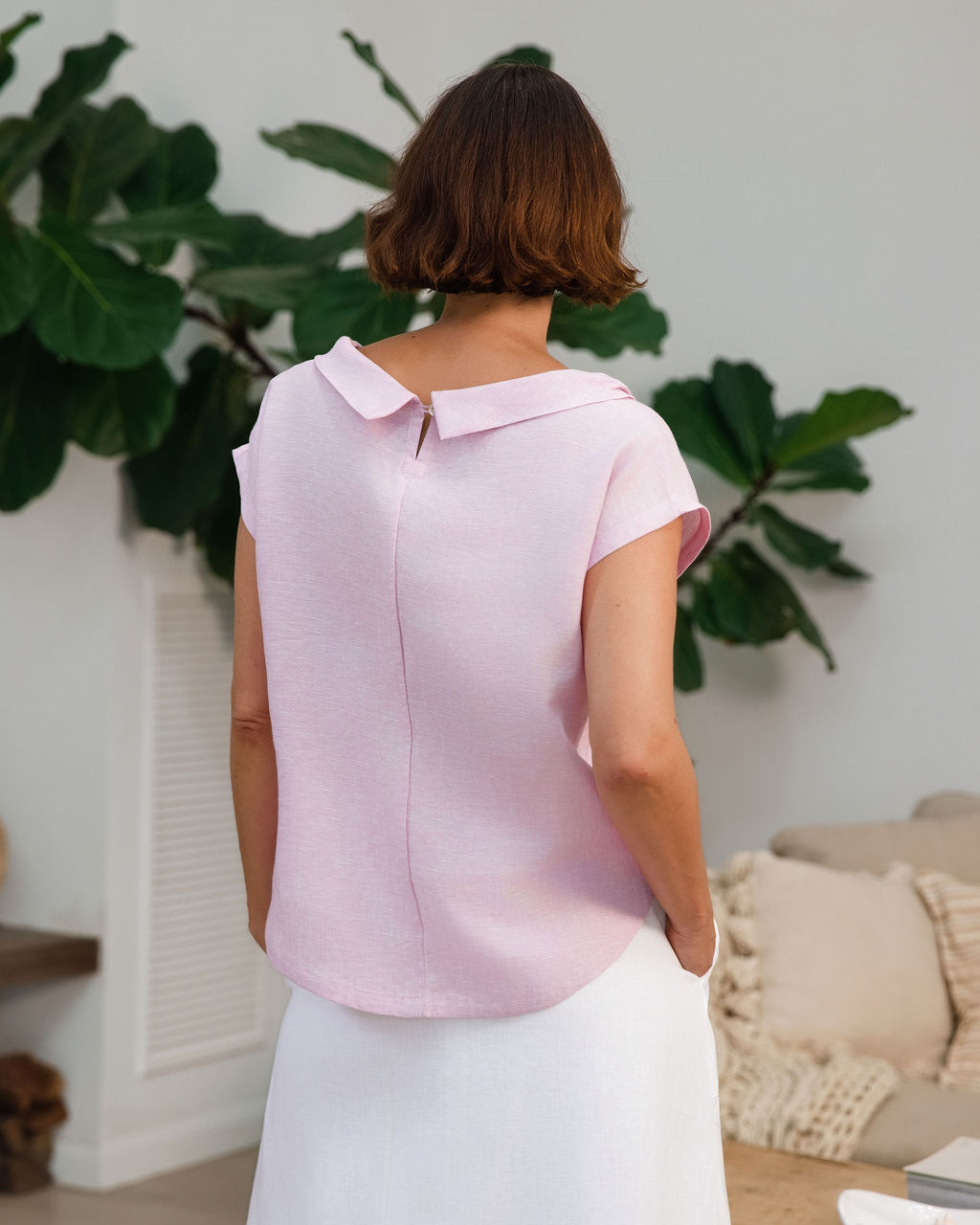 A woman modeling a light pink See Saw linen sleeveless top and white skirt set in a bright, modern living room with a wooden coffee table, sofa, and potted plants.