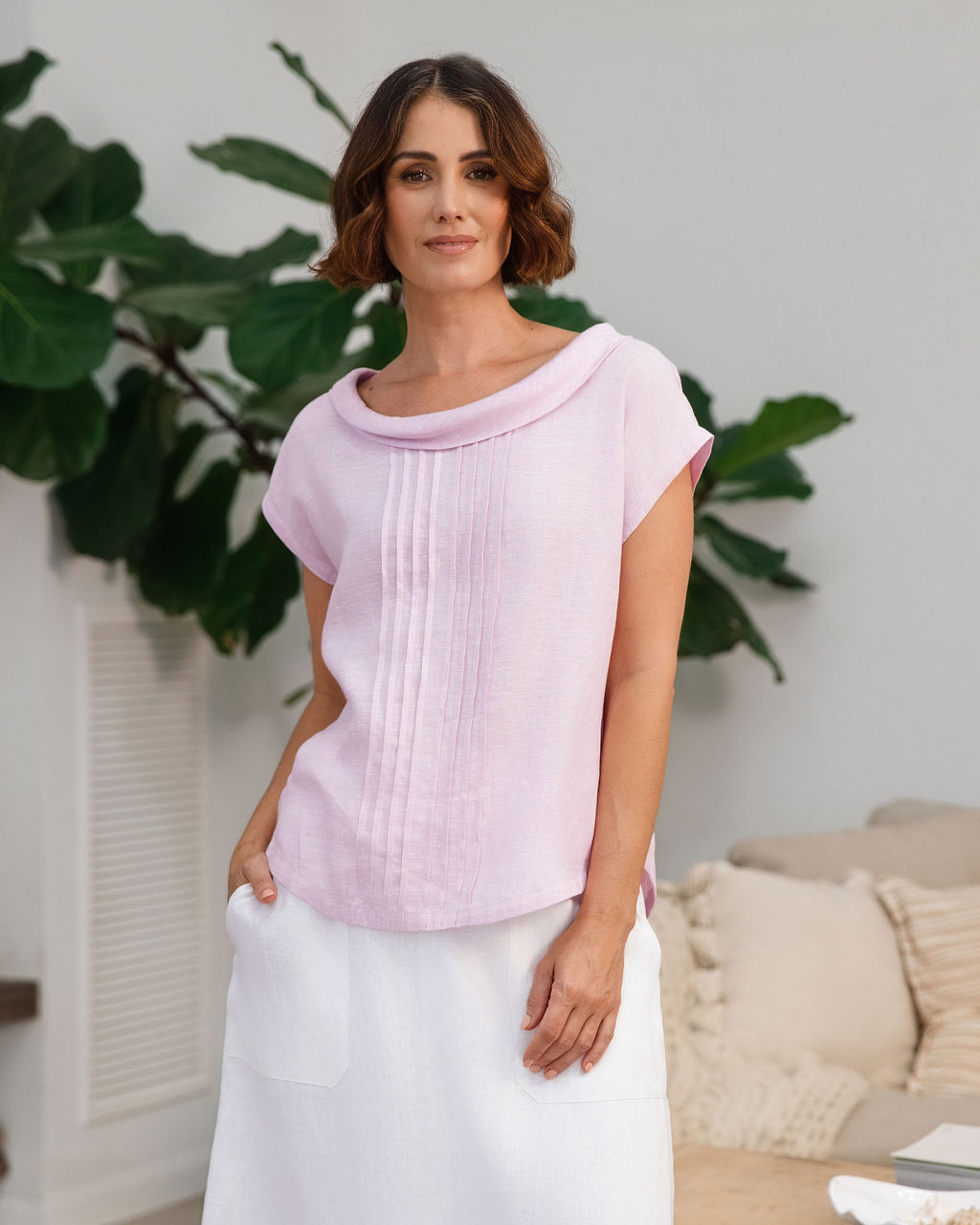A woman modeling a light pink See Saw linen sleeveless top and white skirt set in a bright, modern living room with a wooden coffee table, sofa, and potted plants.