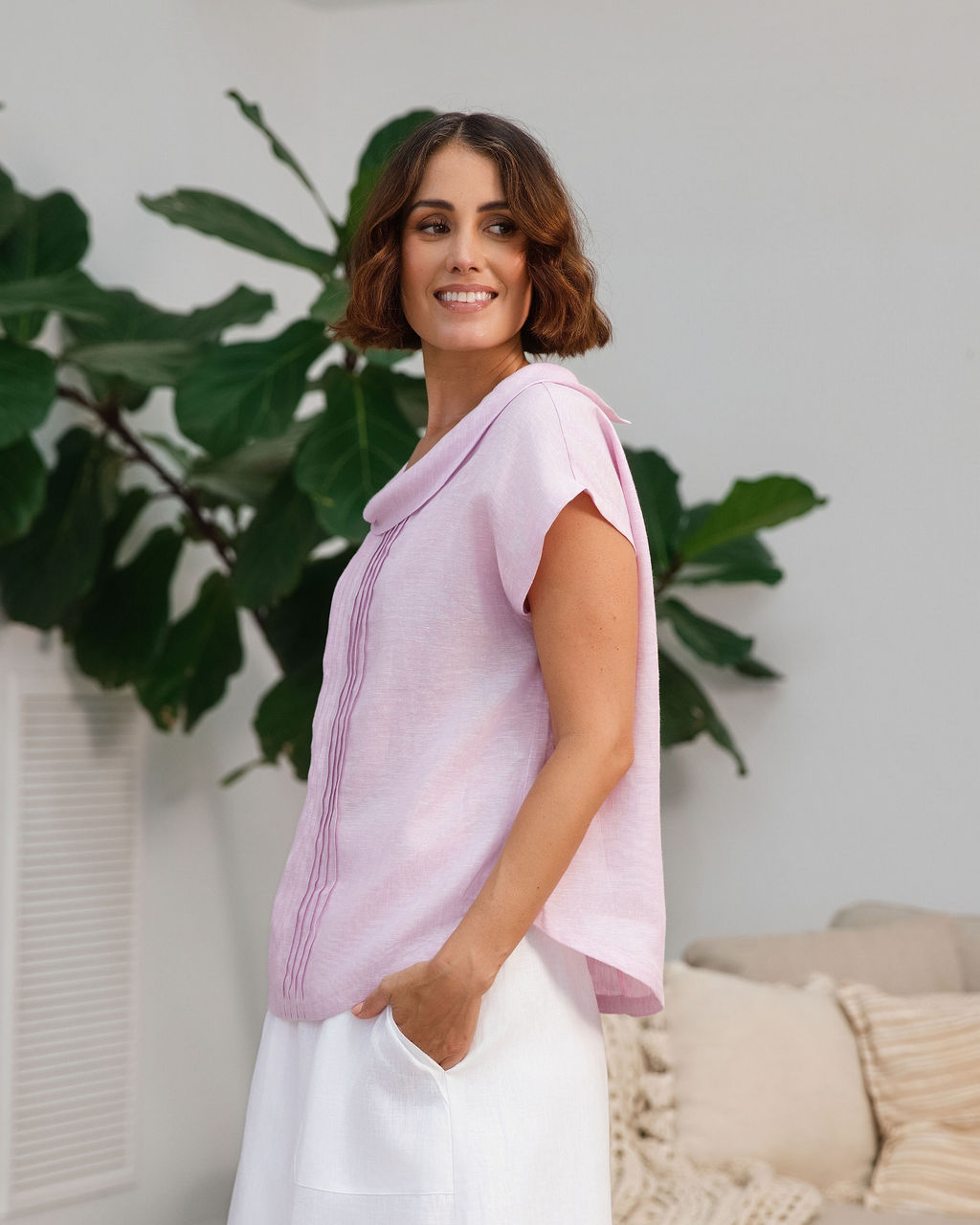 A woman modeling a light pink See Saw linen sleeveless top and white skirt set in a bright, modern living room with a wooden coffee table, sofa, and potted plants.