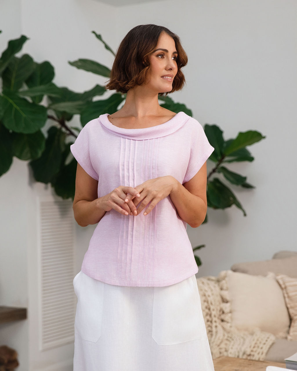 A woman modeling a light pink See Saw linen sleeveless top and white skirt set in a bright, modern living room with a wooden coffee table, sofa, and potted plants.