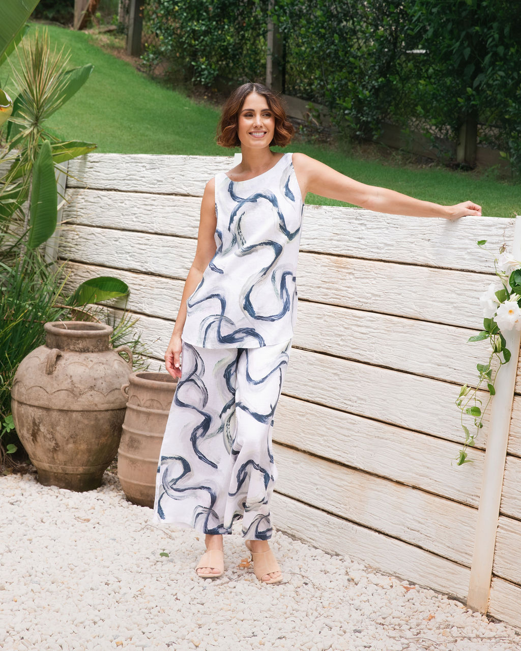 A woman wearing a Riverstone sleeveless white top and matching See Saw wide-leg pants with an abstract blue and gray brushstroke pattern, standing outdoors in a garden setting with large green plants, terracotta pots, and a light stone wall.