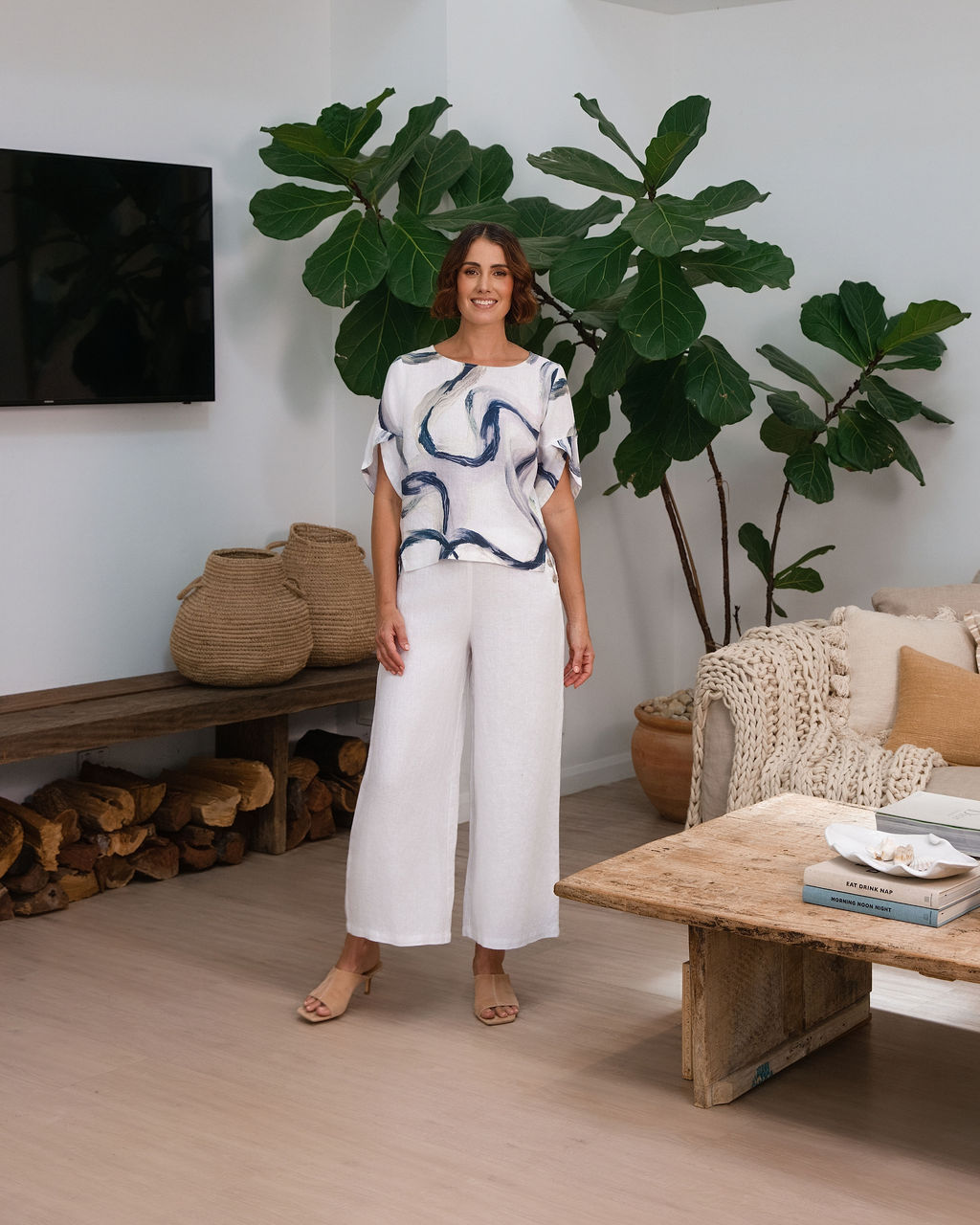 A woman wearing a white See Saw short-sleeve blouse with a blue riverstone brushstroke pattern, paired with white pants, standing in front of a potted fiddle-leaf fig plant.