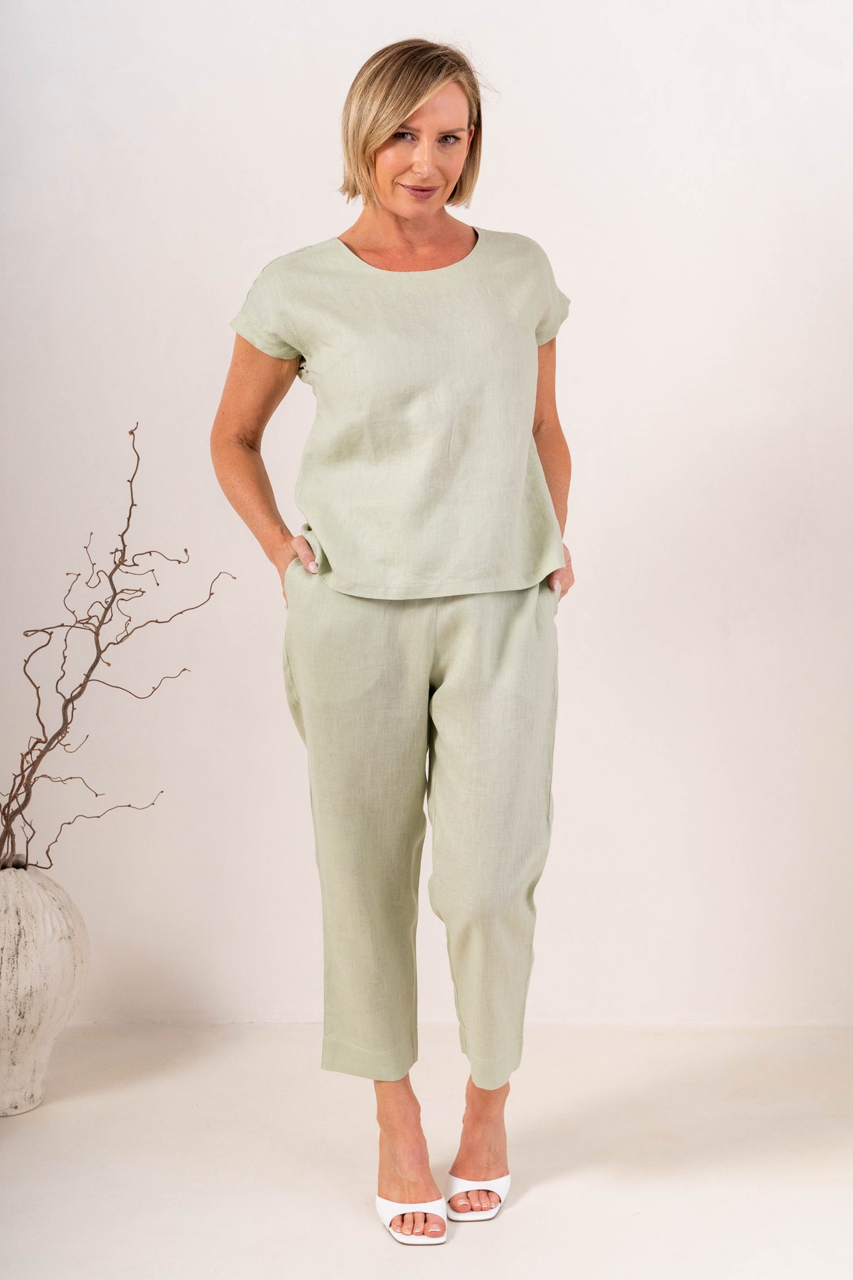 A woman wearing a light sage green See Saw linen short-sleeve top with a button-down back and matching pants, set against a plain off-white background.