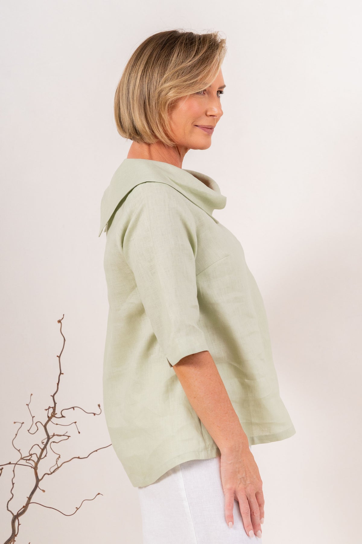 A woman modeling a sage linen See Saw top with a wide draped collar and elbow-length sleeves, paired with white pants, against a plain off-white background.