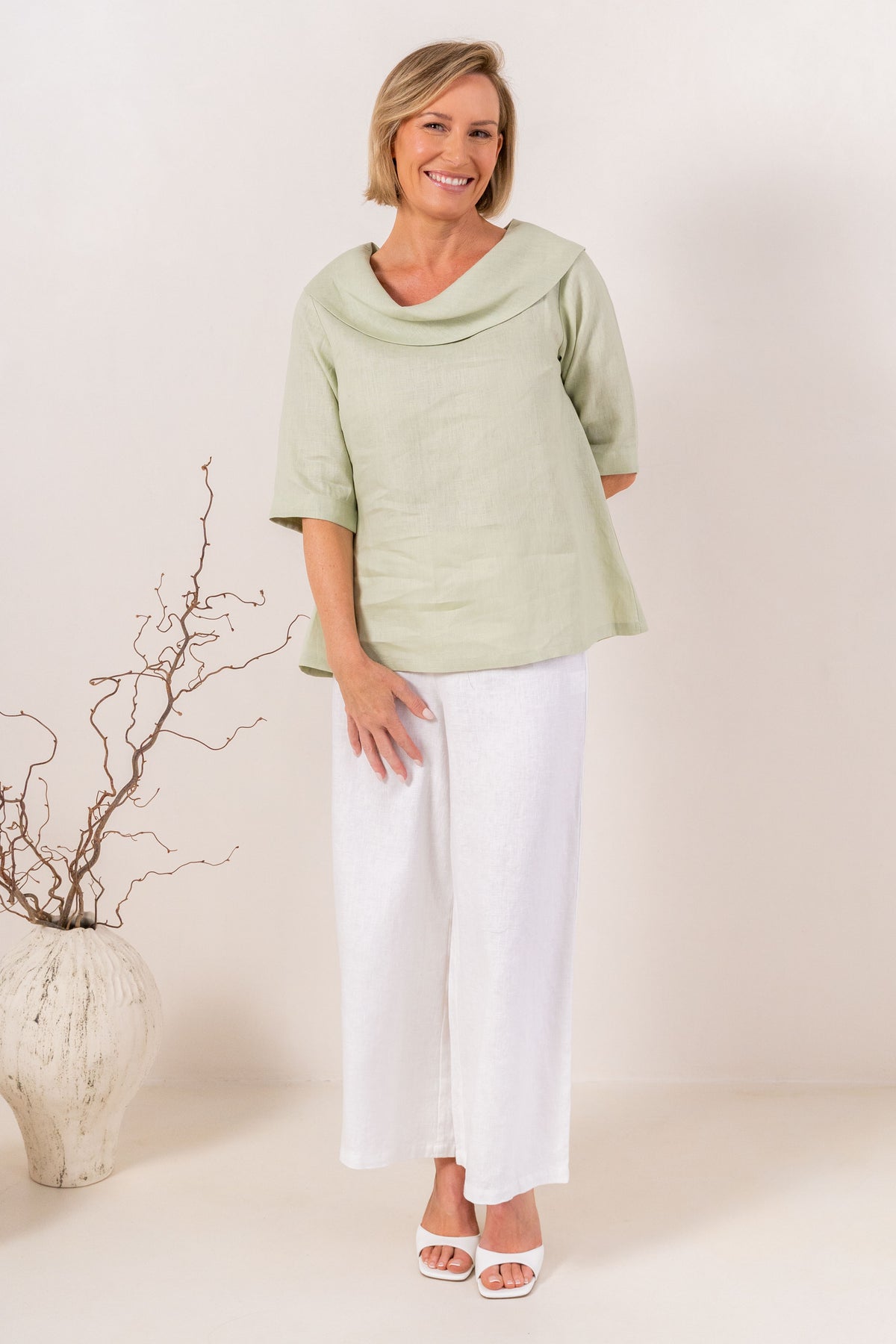 A woman modeling a sage linen See Saw top with a wide draped collar and elbow-length sleeves, paired with white pants, against a plain off-white background.