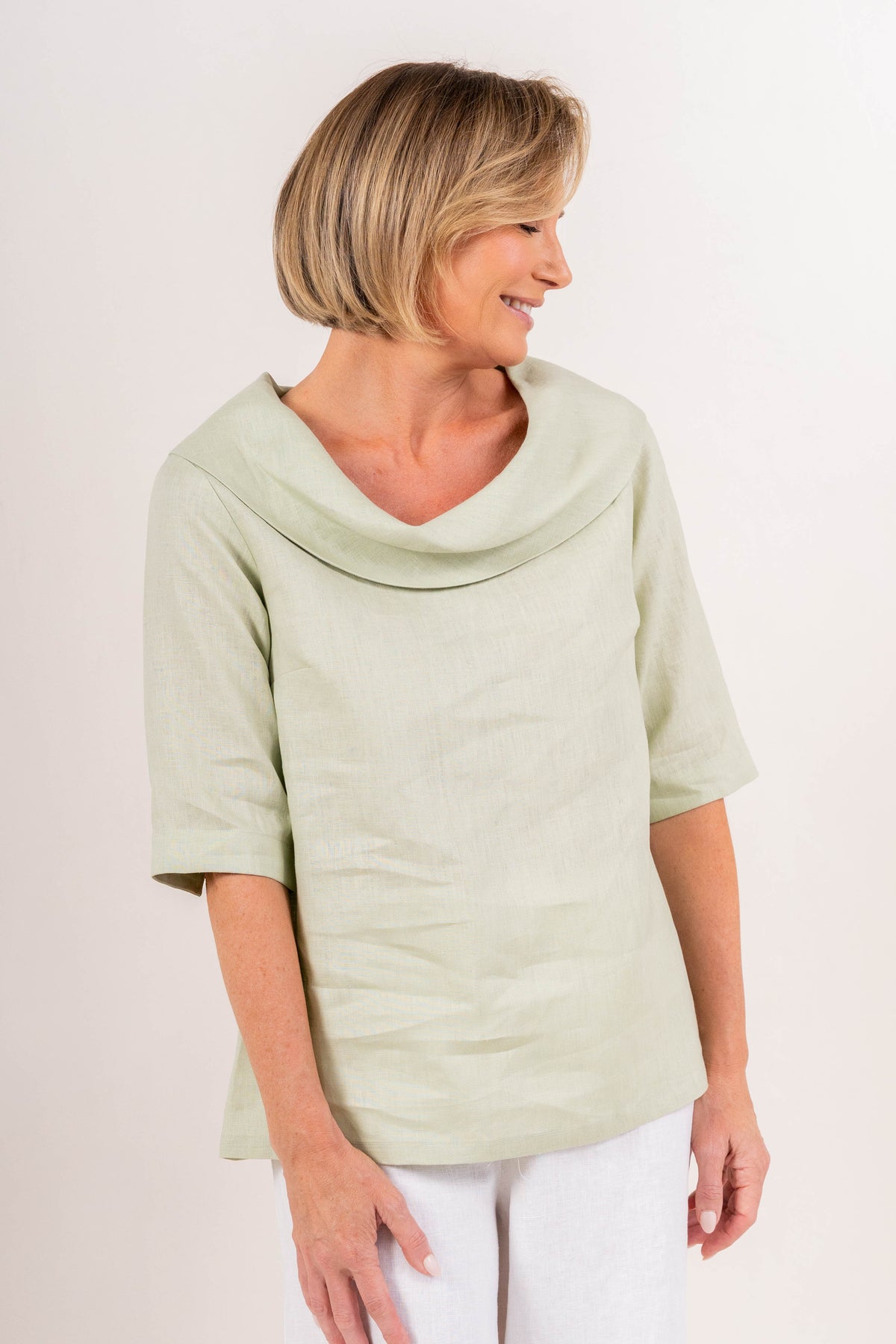 A woman modeling a sage linen See Saw top with a wide draped collar and elbow-length sleeves, paired with white pants, against a plain off-white background.