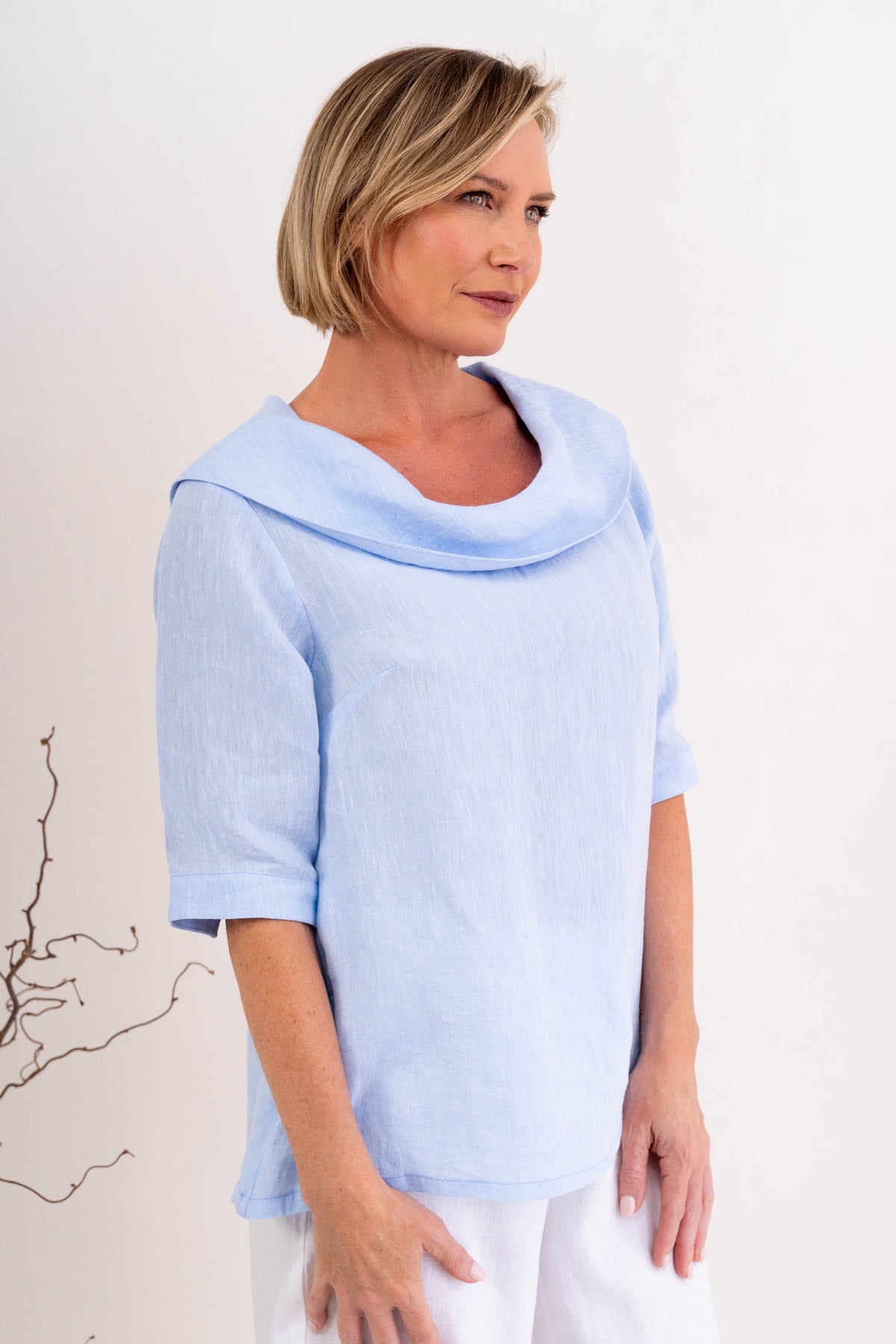 A woman modeling a sky blue linen See Saw top with a wide draped collar and elbow-length sleeves, paired with white pants, against a plain off-white background.