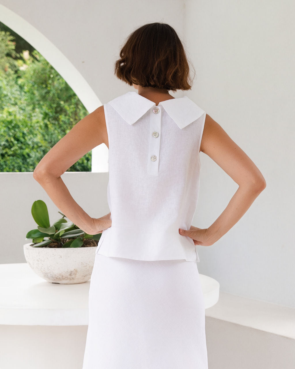 A woman shown from behind wearing a white See Saw Linen sleeveless top with a large pointed collar and three buttons, paired with a white skirt, in a bright white interior setting.