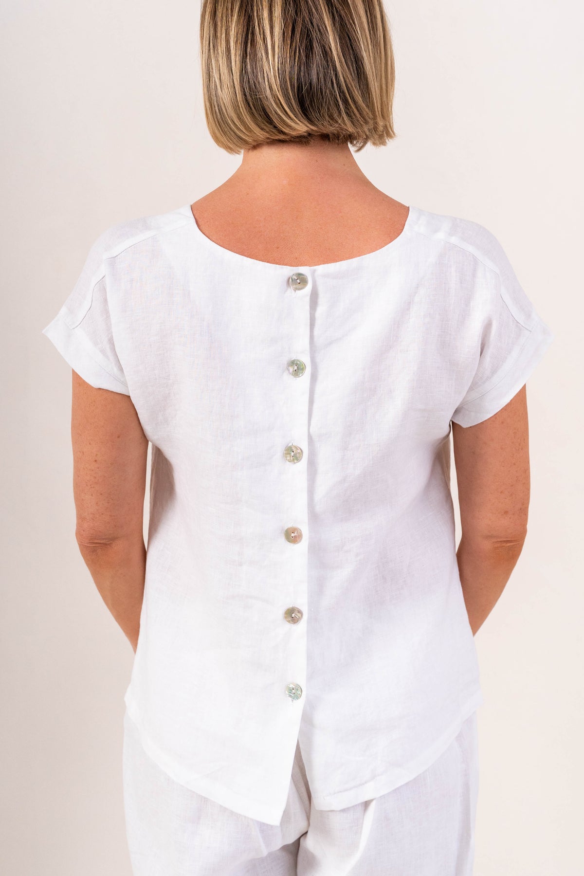 A woman wearing a white short-sleeve See Saw linen top and matching white pants, photographed against a plain light beige studio background.