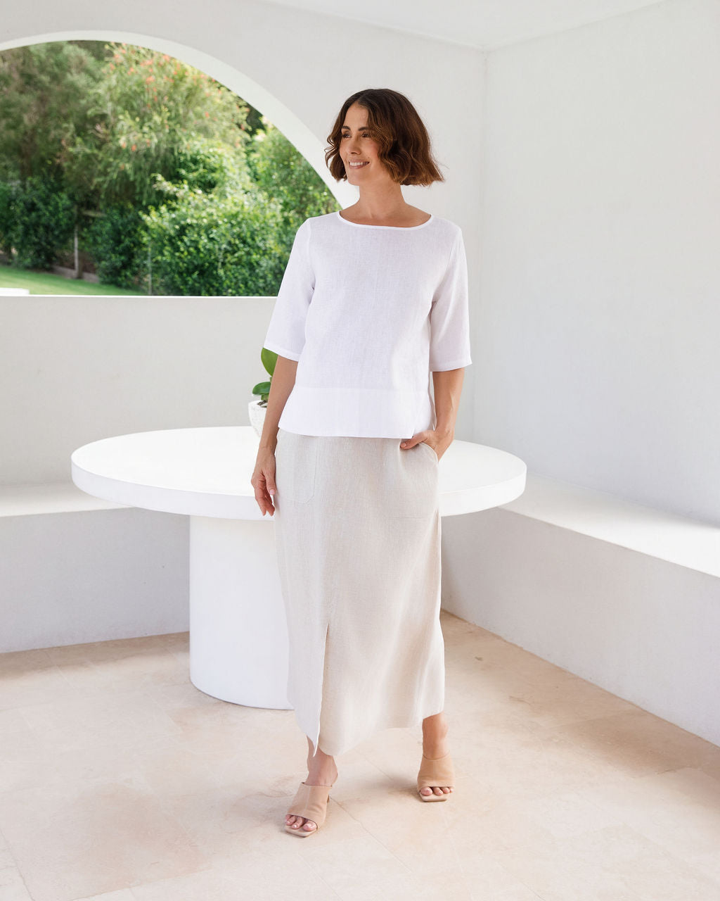 A woman modeling a white linen See Saw three-quarter sleeve top and light beige skirt, standing in a bright, modern white room with a round white table.