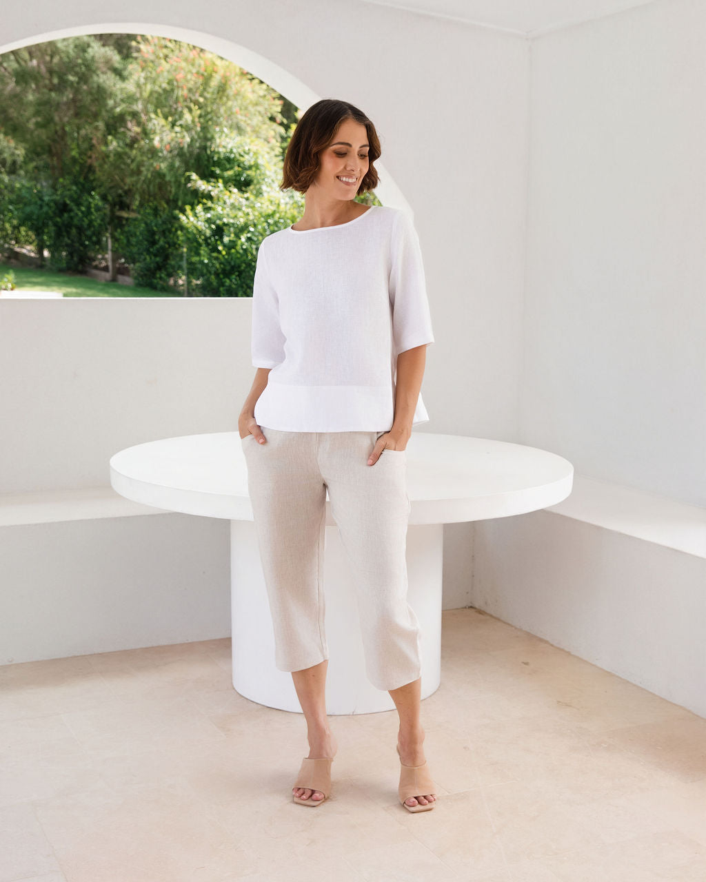 A woman modeling a See Saw linen white three-quarter sleeve top and light beige cropped trousers, standing in a bright, minimalist white room with a round white table.