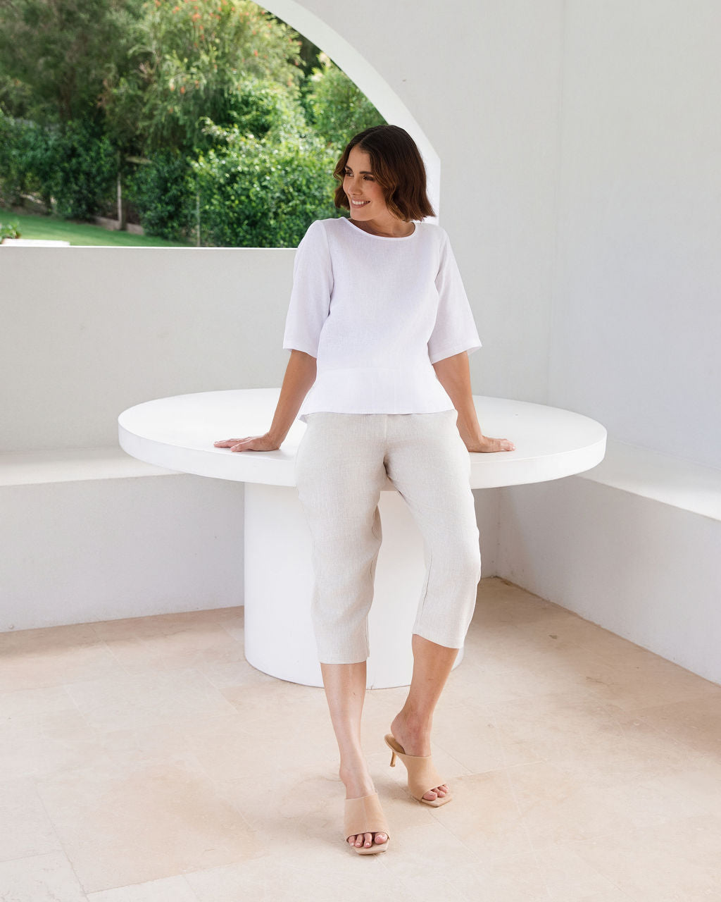 A woman modeling a white linen See Saw three-quarter sleeve top and light beige cropped trousers, standing in a bright, modern white room with a round white table.