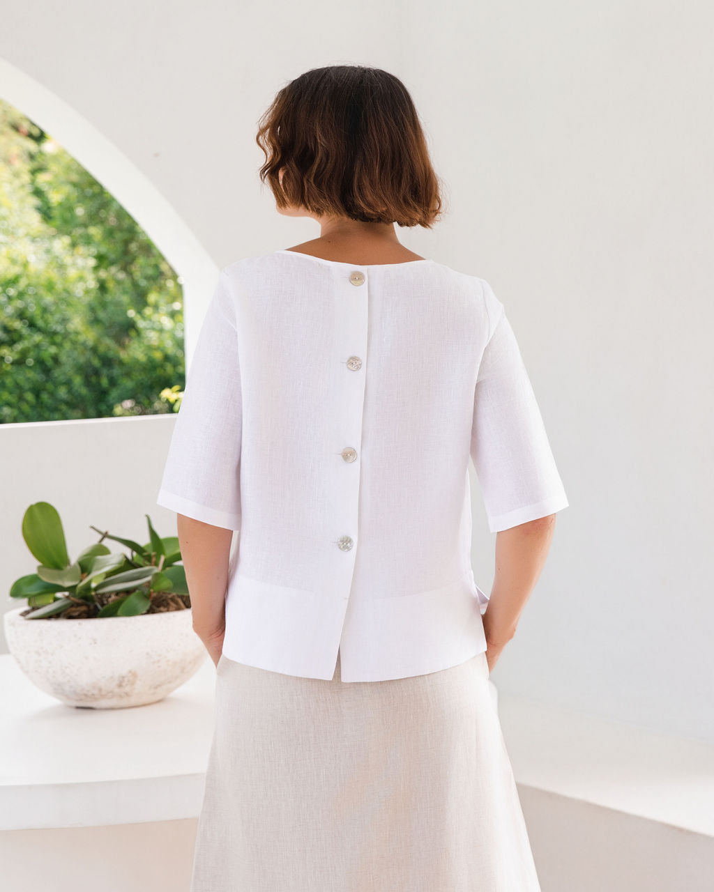 A woman viewed from behind wearing a white short-sleeve blouse with a buttoned back and a light beige skirt, standing in a bright white interior with an arched window and a potted plant visible.