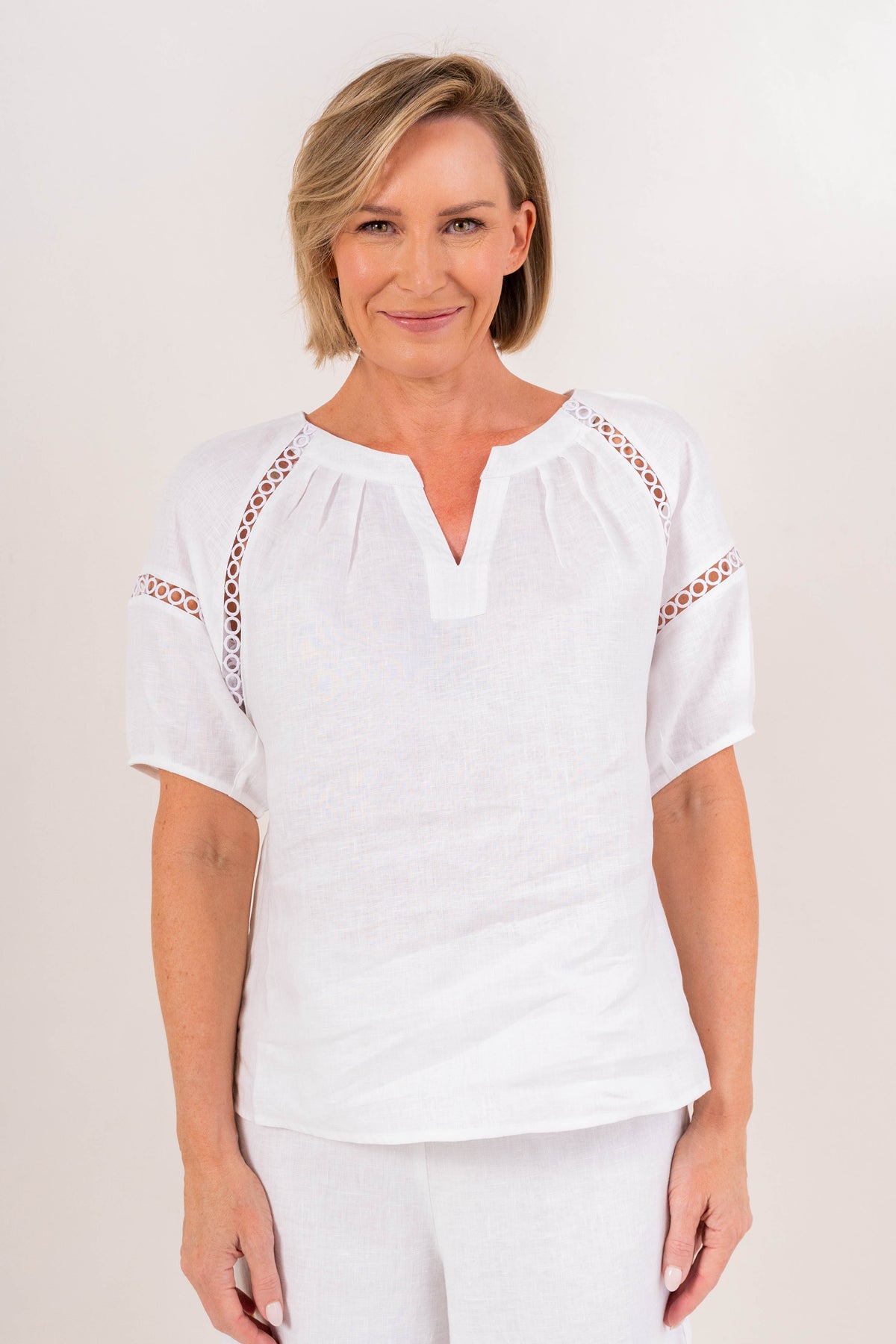 A woman modeling a white short-sleeve linen See Saw top with a split V-neck and decorative openwork lace trim along the shoulders, paired with white pants against a plain light background.