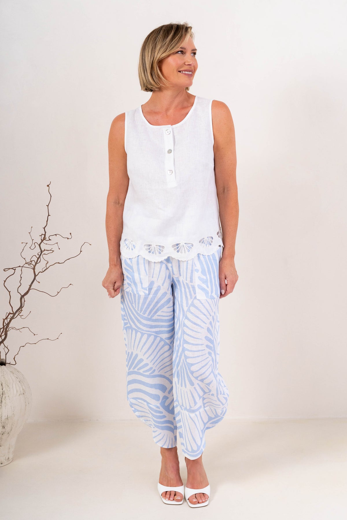 A woman modeling a light blue and white Palm Breze See Saw Linen two-piece set consisting of a short-sleeve top and matching pants, standing against a plain off-white studio background.