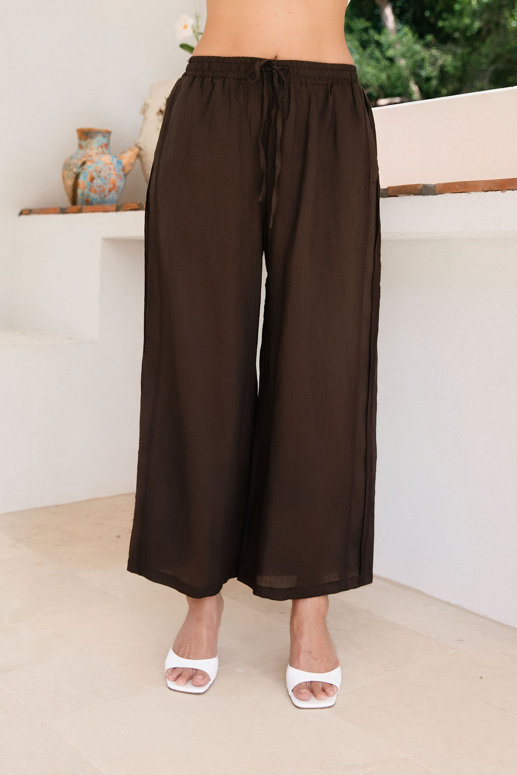 A person wearing See Saw espresso Ramie wide-leg drawstring pants and white heeled sandals, standing indoors against a light neutral wall.