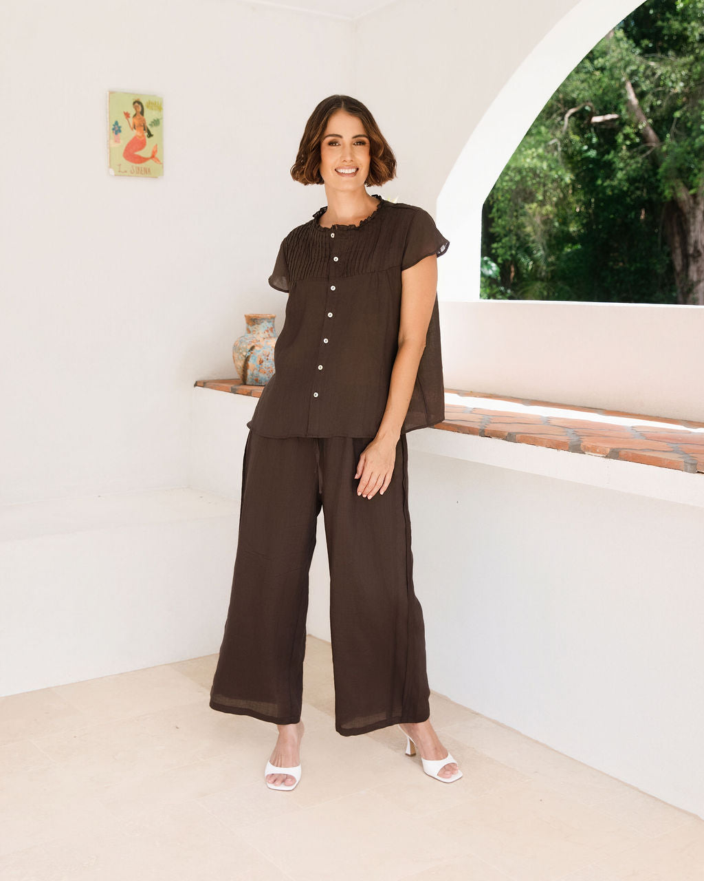 A woman modeling a dark espresso See Saw Ramie two-piece outfit consisting of a short-sleeve button-front top with draped sleeves and matching wide-leg pants, standing in a bright white architectural setting.