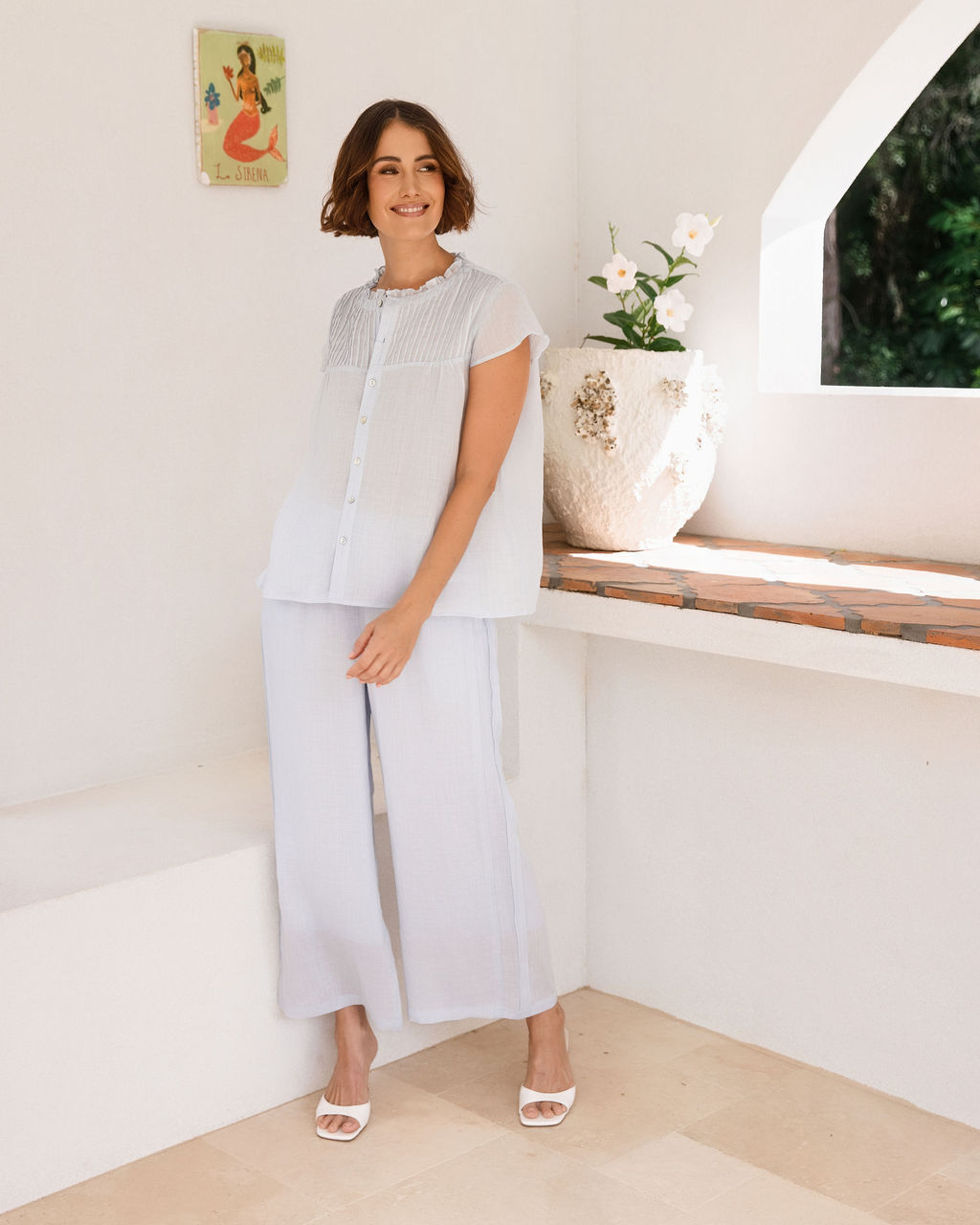 A woman modeling a See Saw Ramie blue mist short-sleeve button-front top and matching white wide-leg cropped pants, standing in a bright white architectural interior with an arched window showing greenery outside.