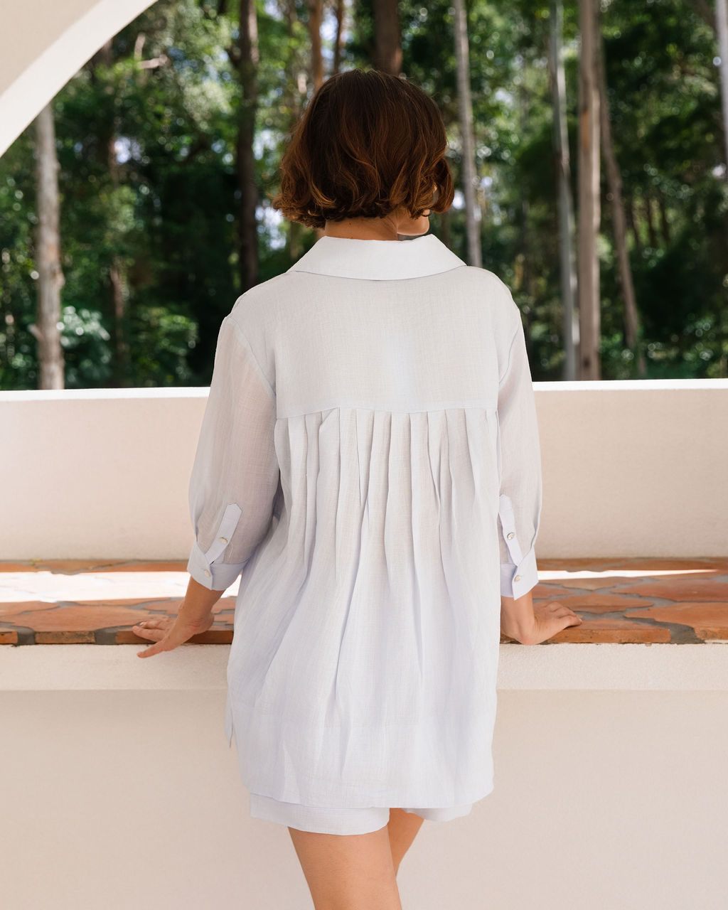 A woman wearing a light blue long-sleeve button-up See Saw Ramie shirt and matching shorts, standing in a bright, modern white architectural setting with a tiled bench and greenery in the background.
