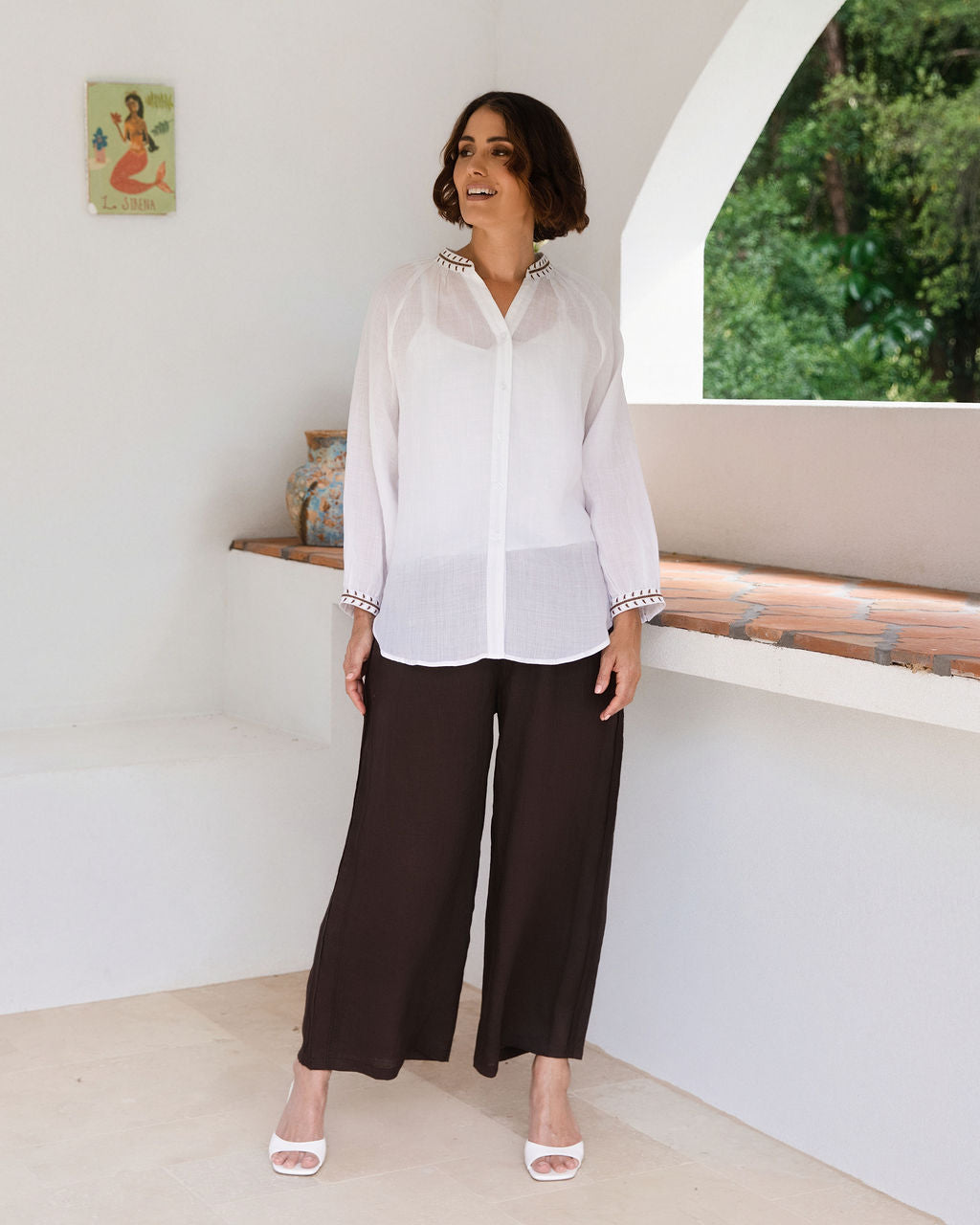 A woman modeling a white See Saw Ramie button-front top and espresso wide-leg pants, standing in a bright white architectural interior with a tiled ledge and potted plant.