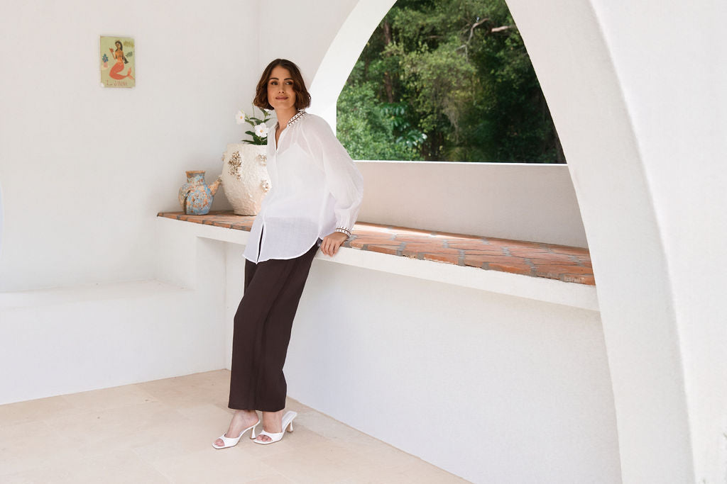A woman modeling a white See Saw Ramie button-front top and espresso wide-leg pants, standing in a bright white architectural interior with a tiled ledge and potted plant.