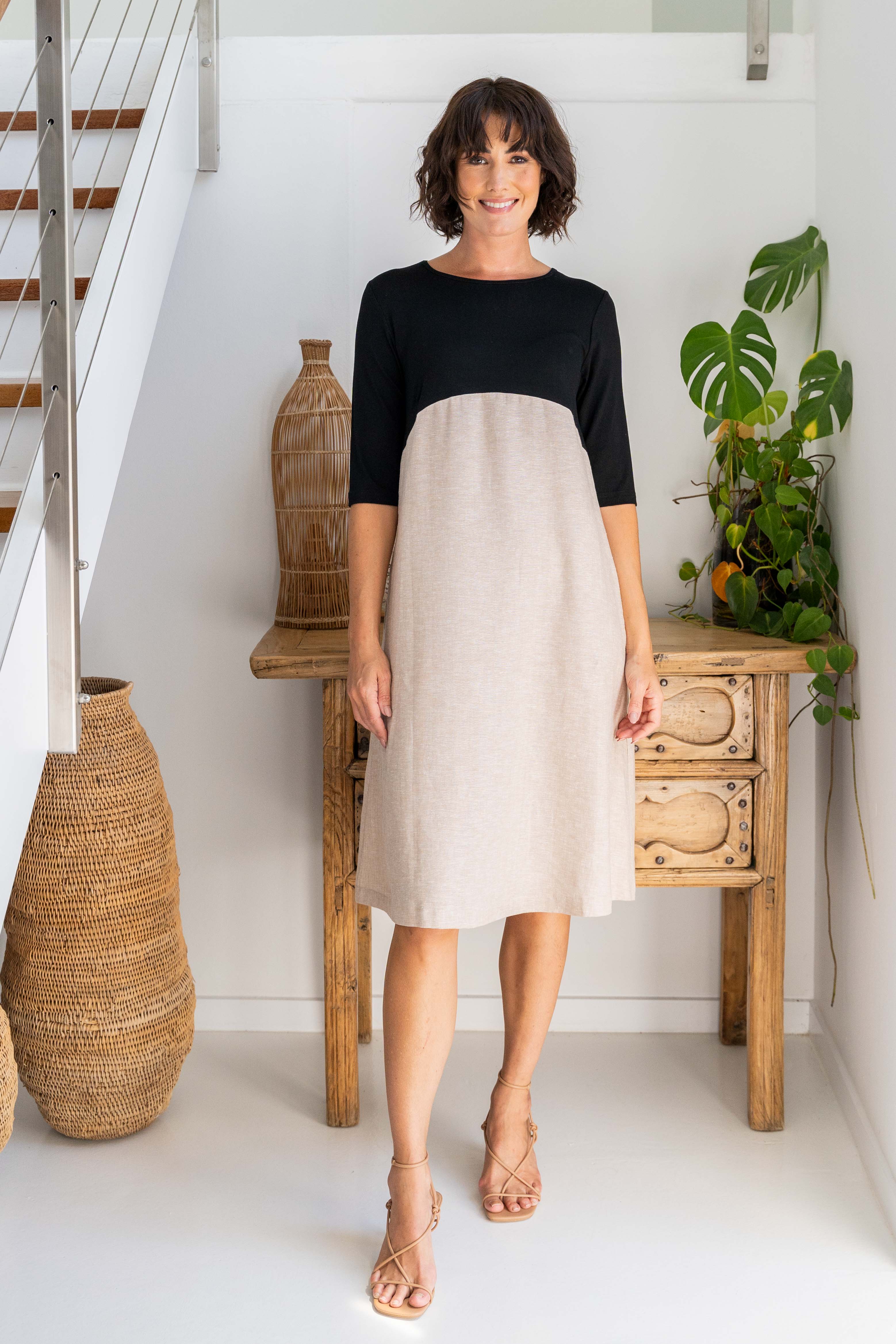 Woman wearing a rayon and linen See Saw dress standing in front of a white background and drawers and a plant.