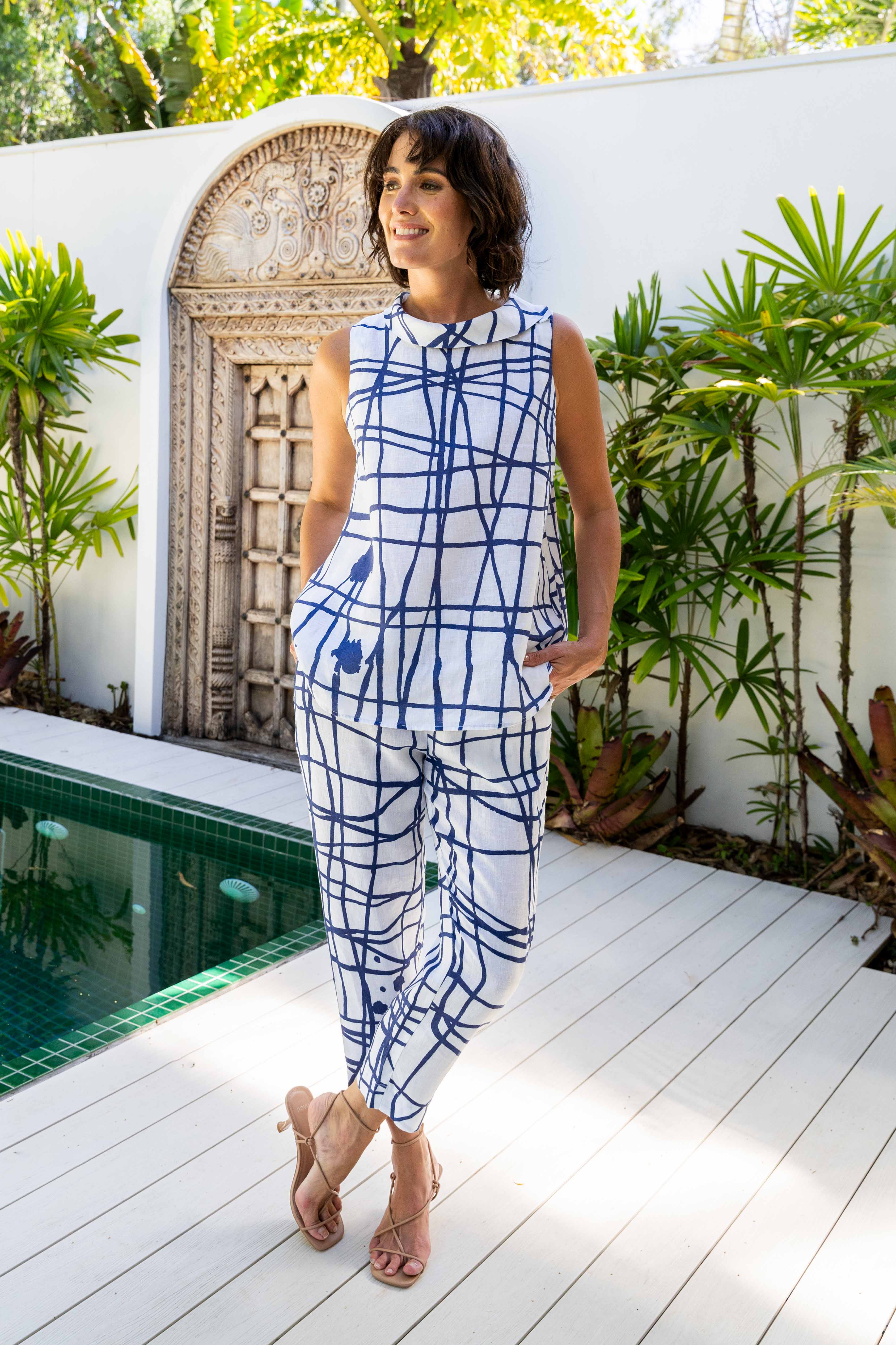 Woman in a blue and white patterned See Saw linen pant outfit standing by a pool with plants and a decorative wall in the background.