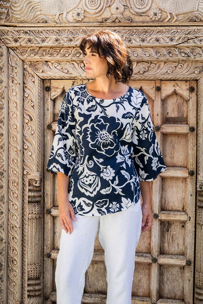 Woman wearing a linen see saw navy floral blouse and white pants standing in front of an ornate wooden door.