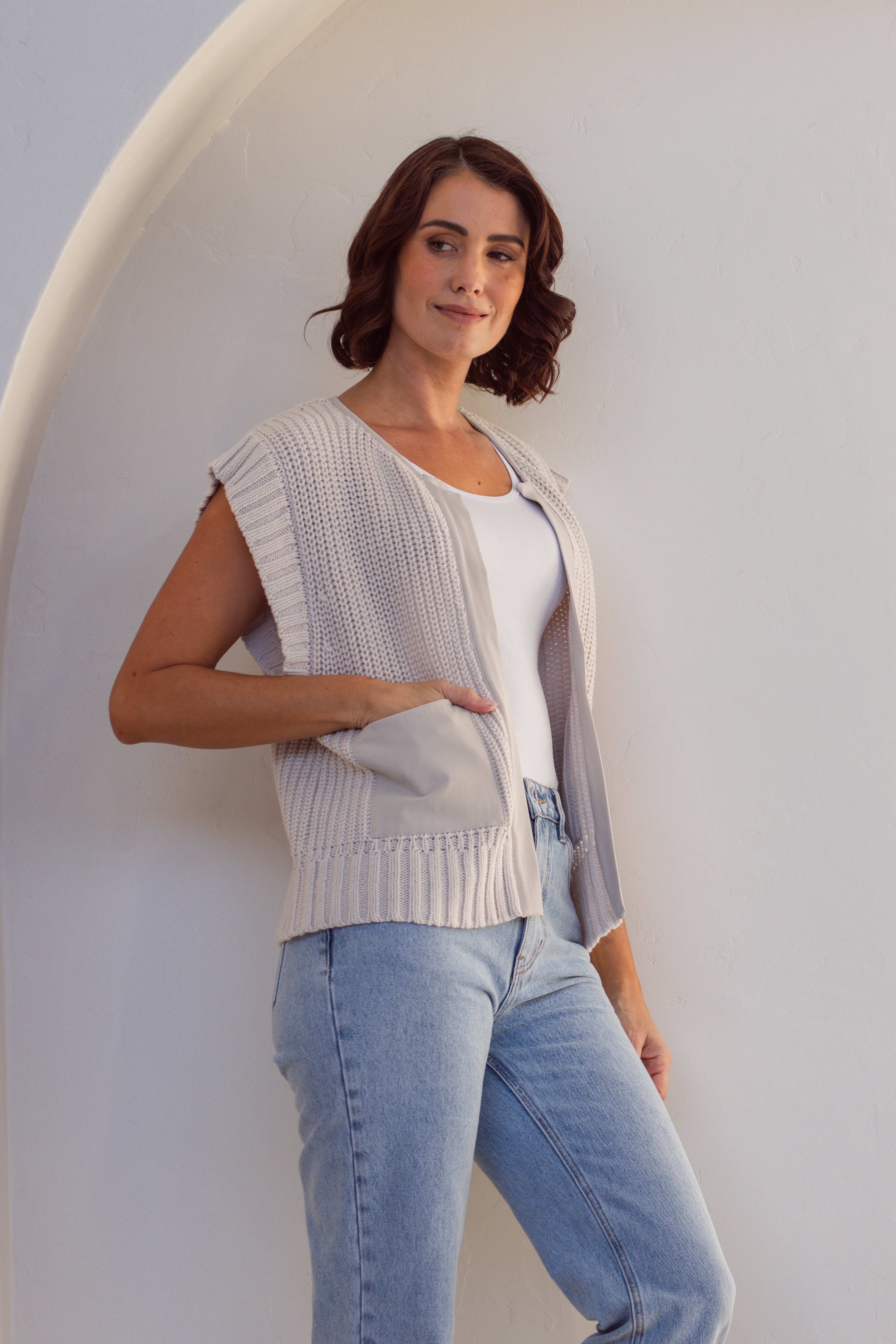 Woman wearing a stone cotton See Saw knitted vest over a white shirt and blue jeans against a white wall.
