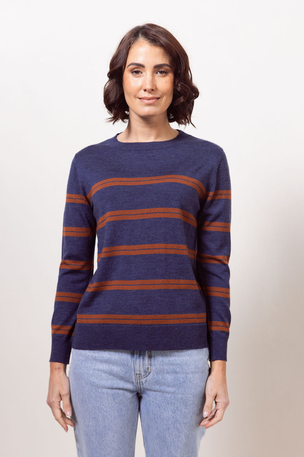 Woman wearing a indigo See Saw Merino wool sweater with copper stripes and dark jeans against a plain background