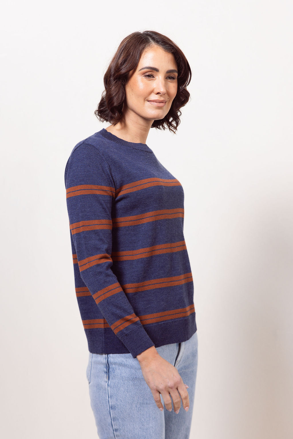 Woman wearing a indigo See Saw Merino wool sweater with copper stripes and dark jeans against a plain background