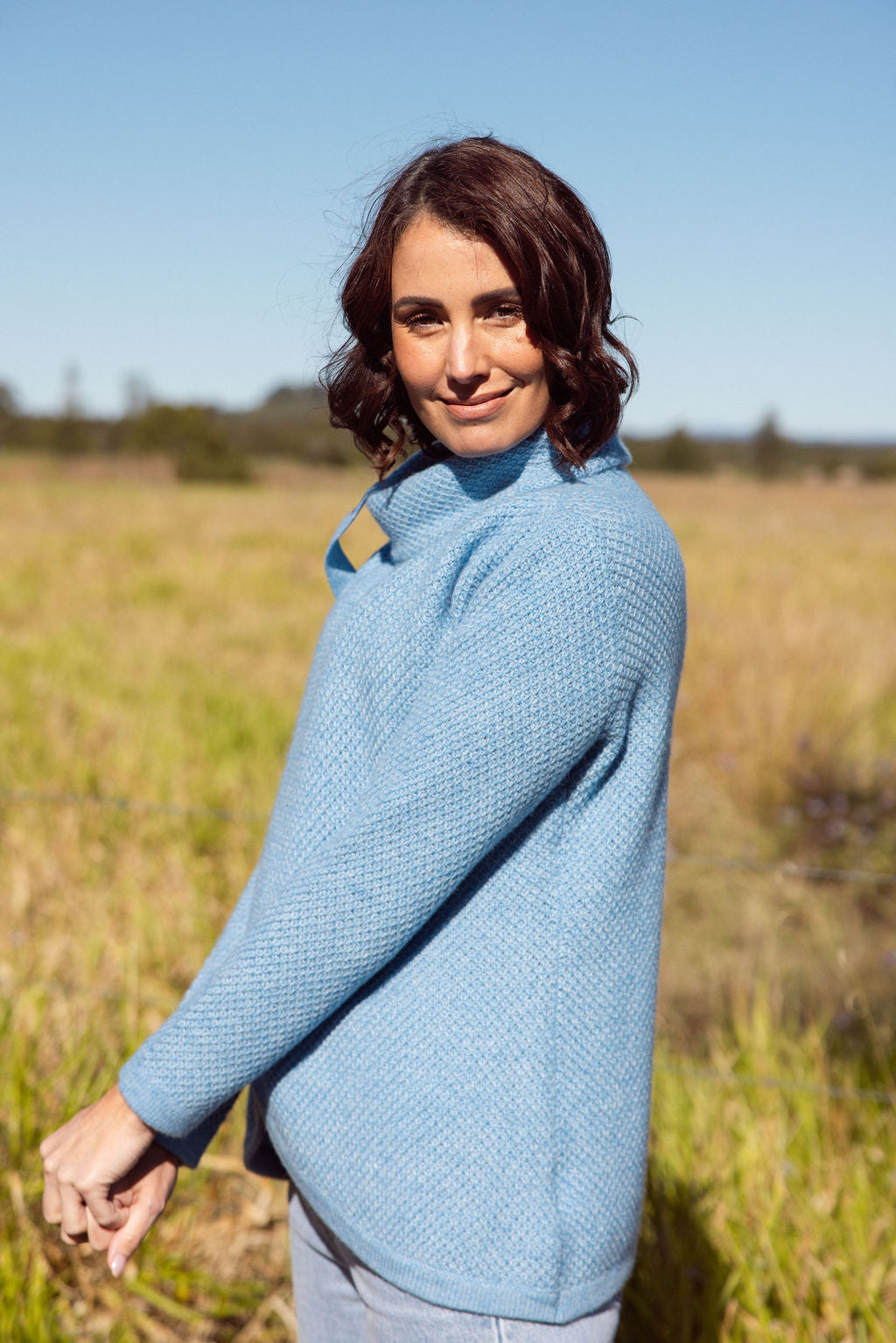 Woman wearing a powder blue Cowl Neck Moss Stitch See Saw sweater with an outdoor background