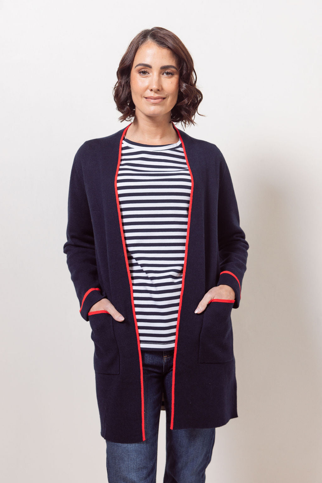 Woman wearing a See Saw navy cardigan with red trim, striped shirt, jeans, and white sneakers on a plain background