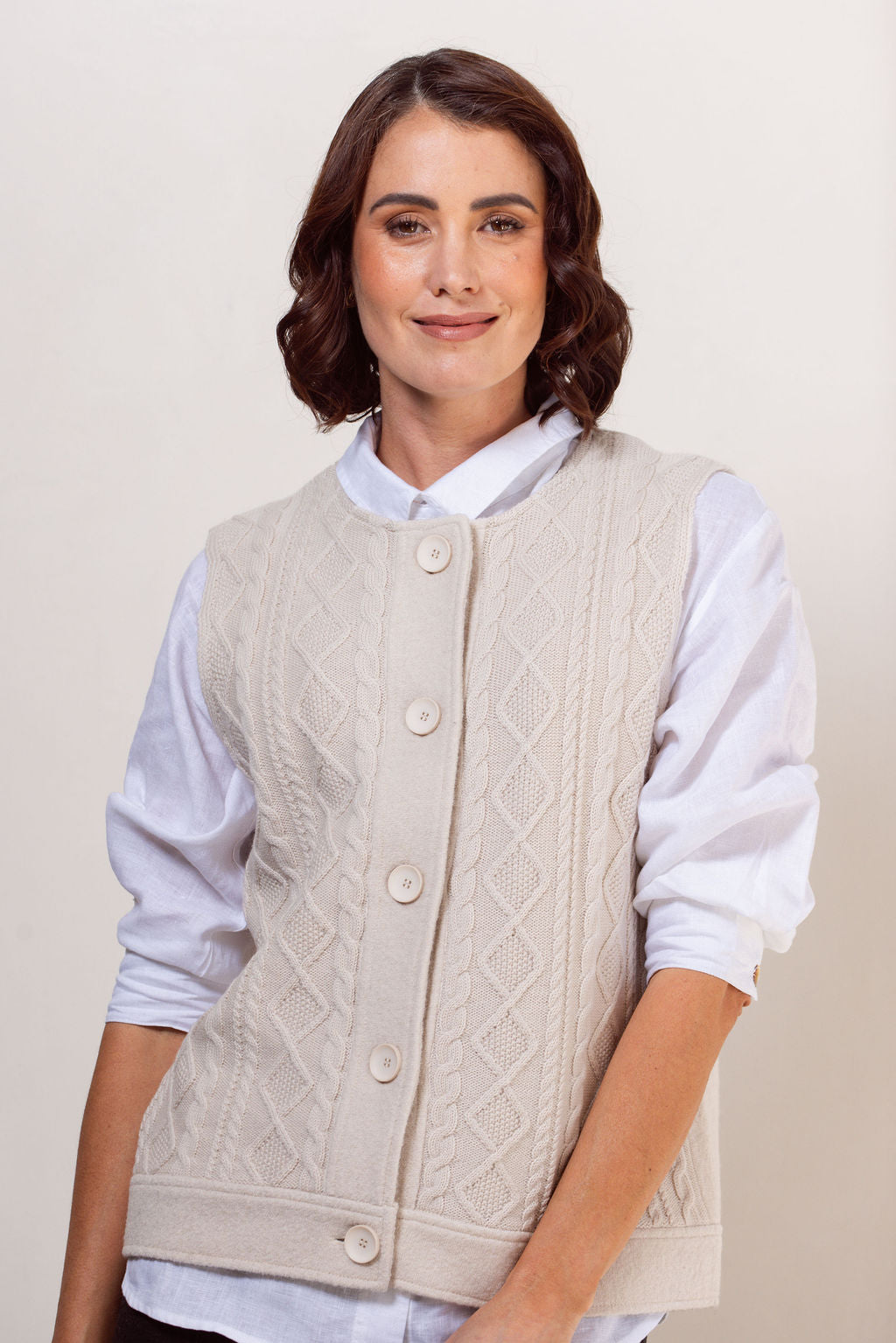 Woman wearing a beige cable knit vest over a white shirt against a plain background