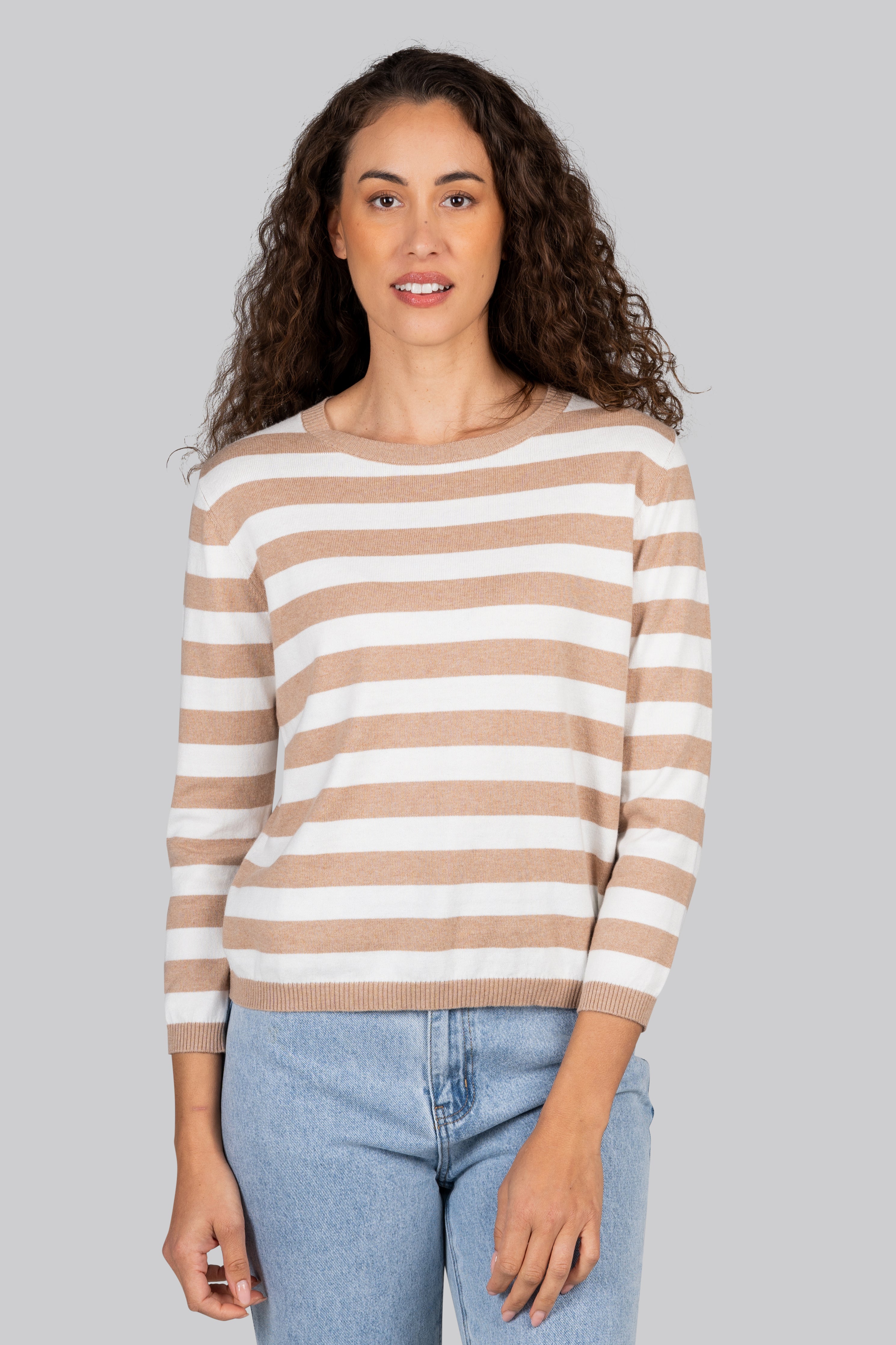 Woman wearing a beige and white striped Kaleido sweater with blue jeans on a gray background