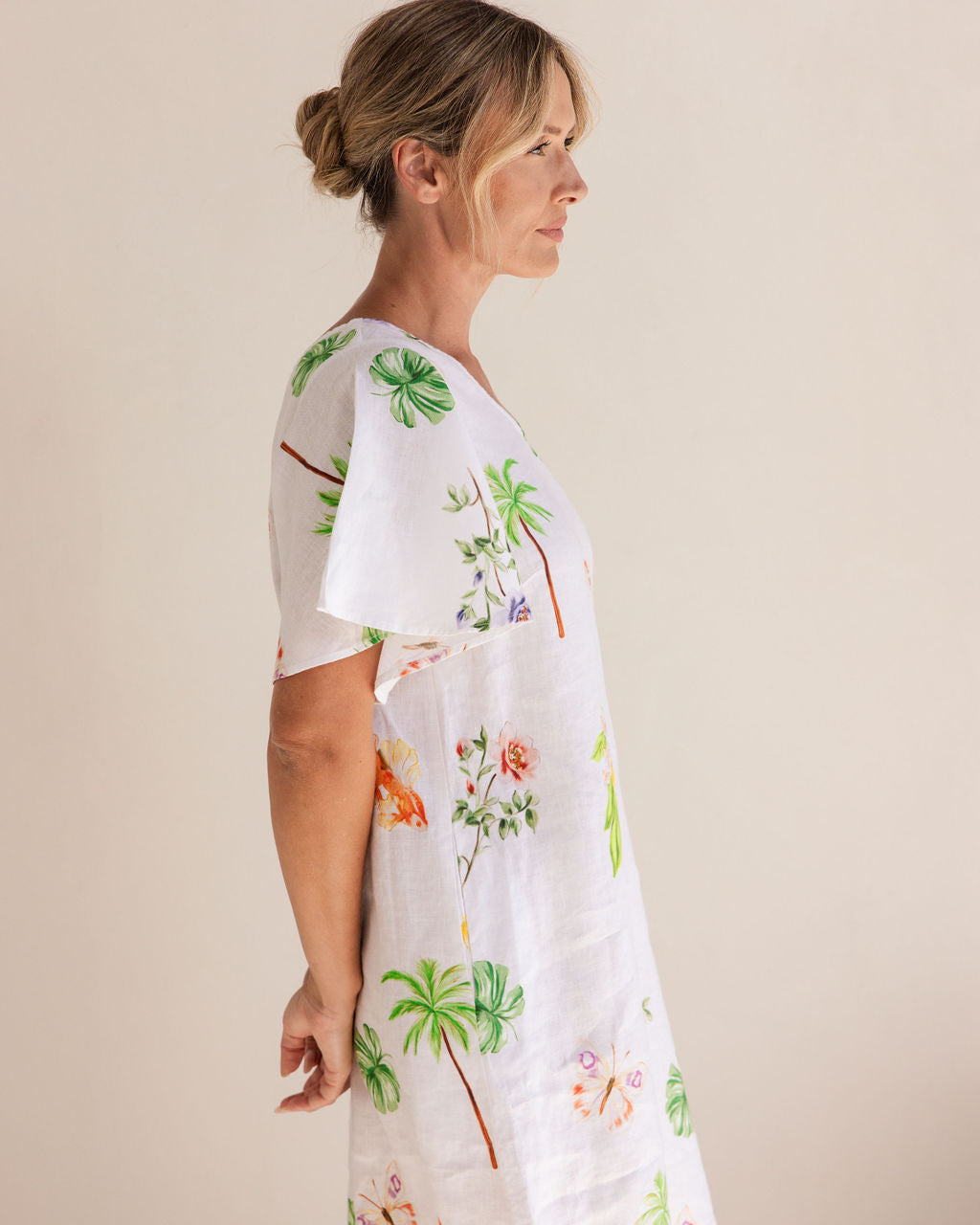 Woman wearing a white dress with tropical floral and palm tree patterns on a beige background. See Saw Linen V Neck Flutter Sleeve Midi Dress