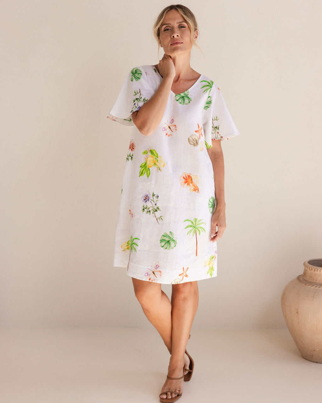 Woman wearing a white dress with colorful floral and palm tree patterns on a beige background. See Saw Linen V Neck Flutter Sleeve Midi Dress