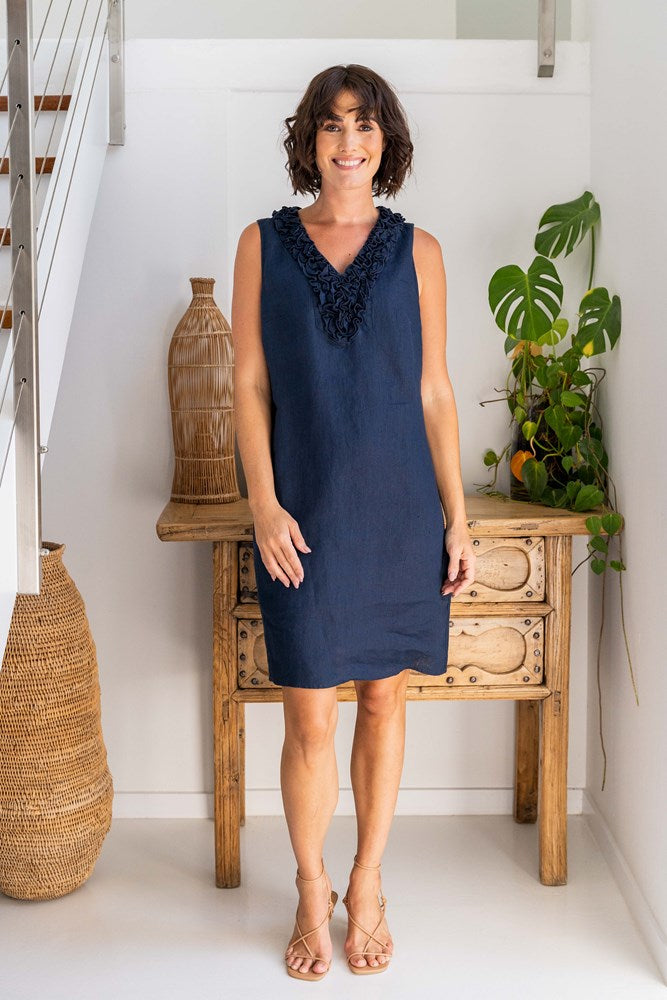 Woman wearing a navy dress standing in a room with a wooden table and decorative items. See Saw Linen Ruffle Front Dress