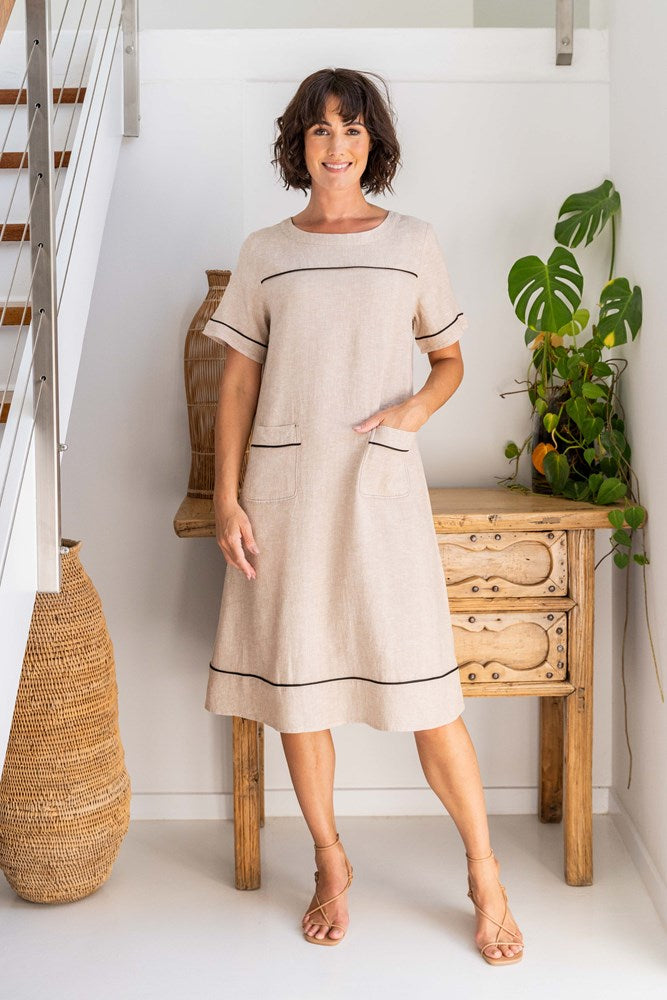Woman wearing a beige dress standing in a room with wooden furniture and plants. See Saw Linen Stitch Detail A line Dress