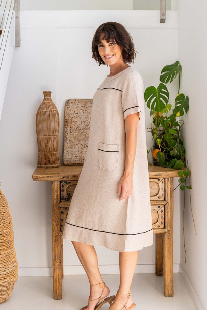 Woman wearing a beige dress with black trim in a room with wooden furniture and plants. See Saw Linen Stitch Detail A line Dress