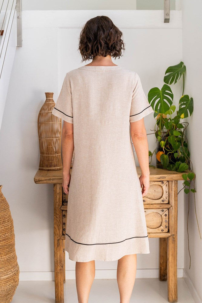Woman wearing a beige dress with black trim standing in a room with a wooden table and plants. See Saw Linen Stitch Detail A line Dress
