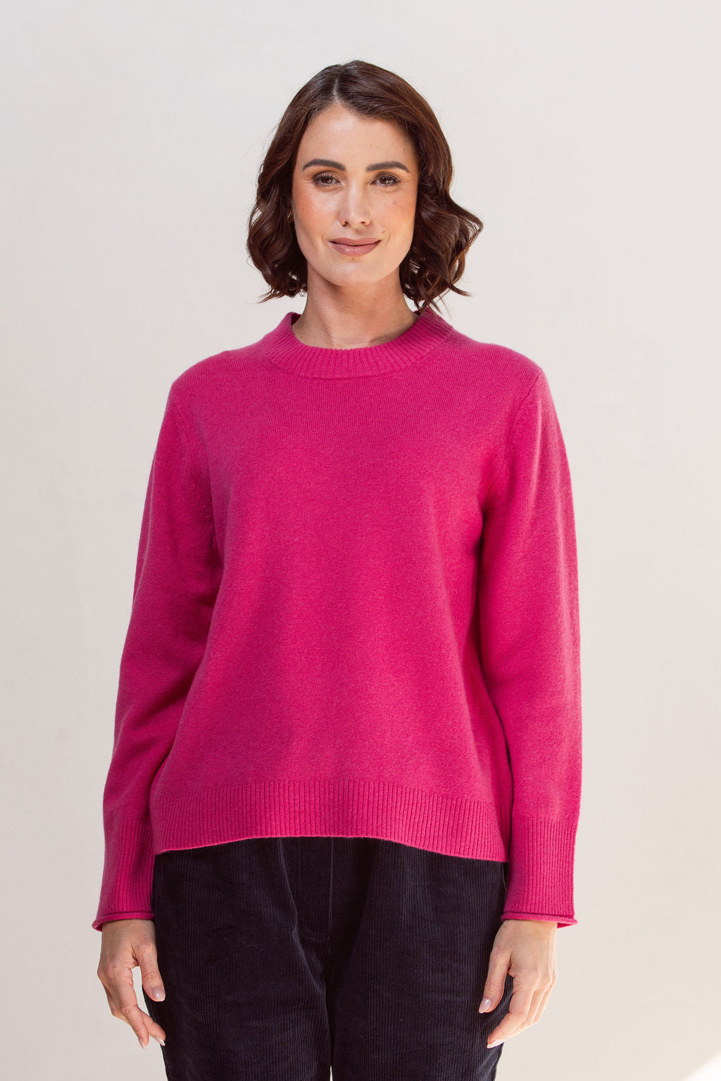Woman wearing a pink crew neck See Saw sweater and light blue jeans against a plain background